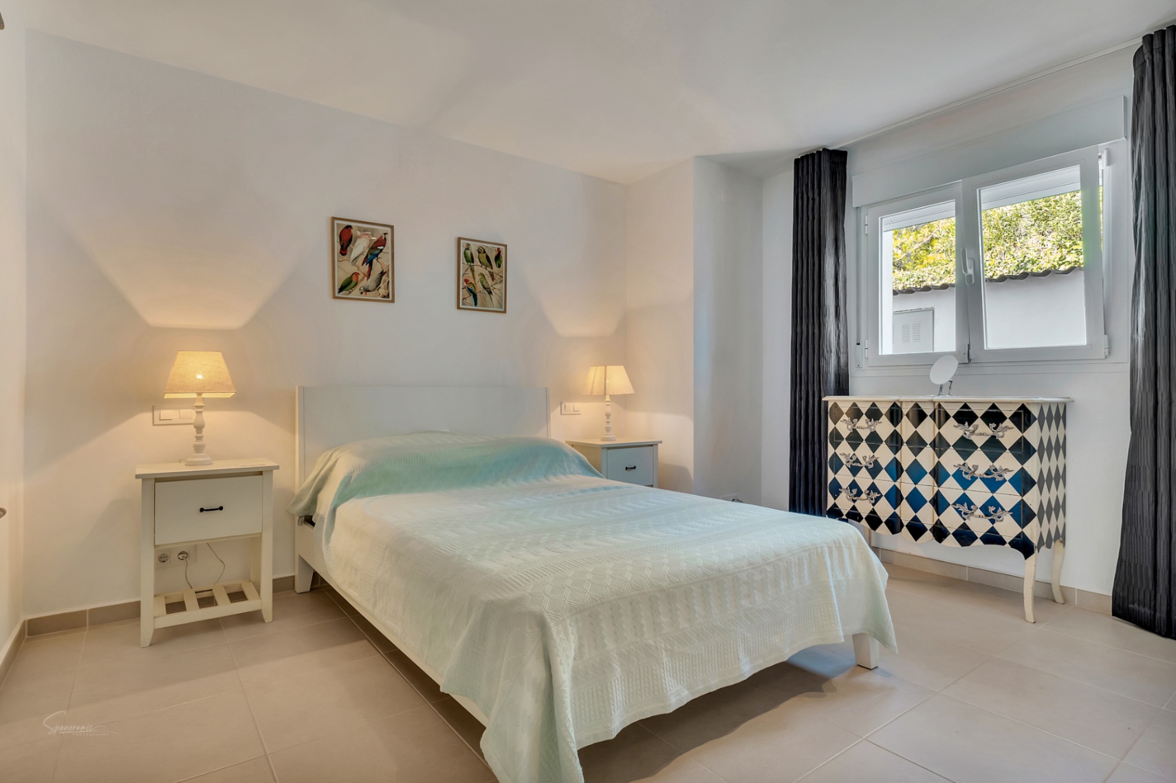 Chambre lumineuse avec sol en grès cérame, lit double, tables de chevet blanches et une commode décorative à motifs de losanges près de la fenêtre.