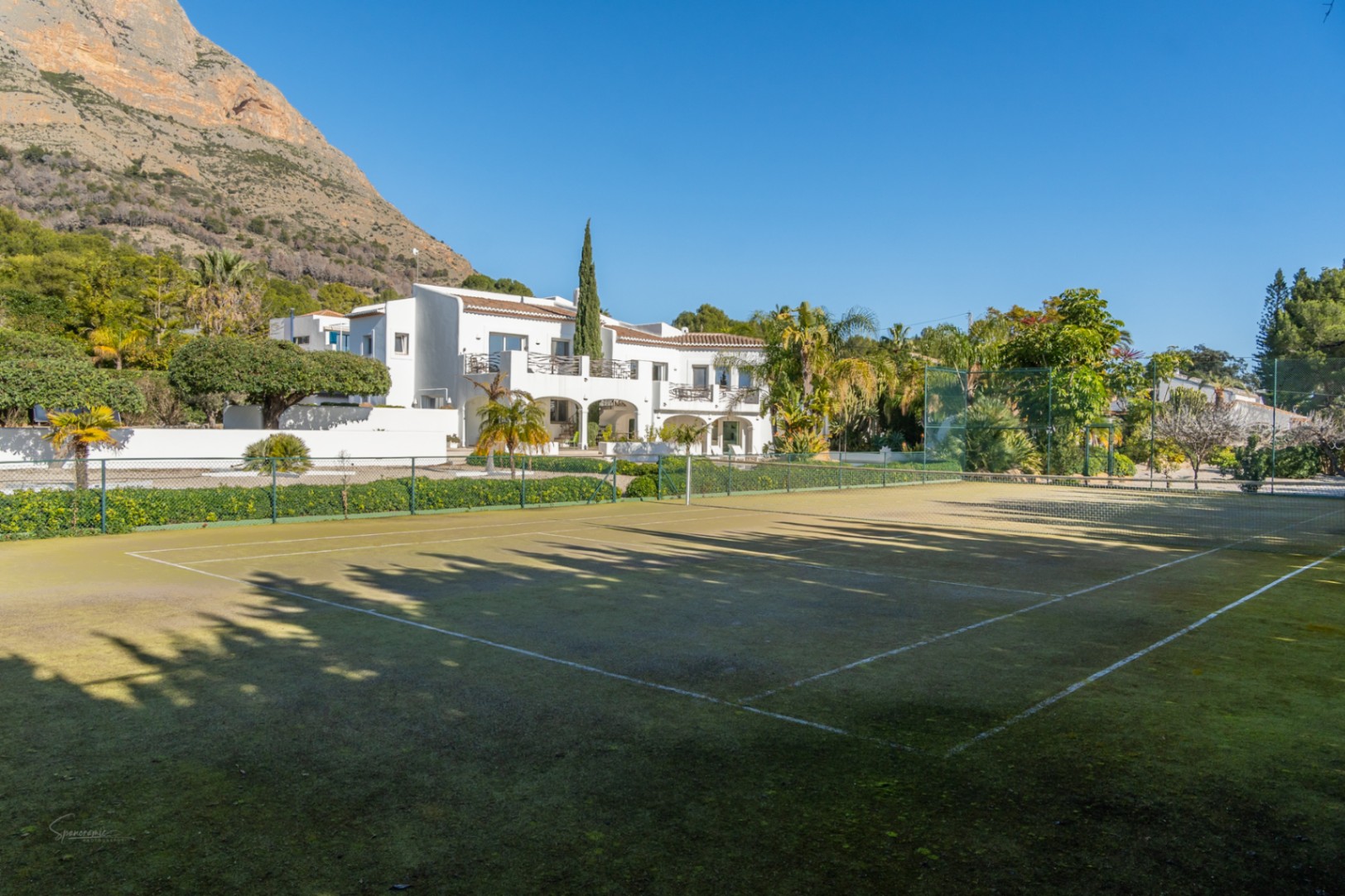 Court de tennis privé avec surface verte et clôture, situé devant la villa de style Ibiza avec le massif du Montgó en arrière-plan.