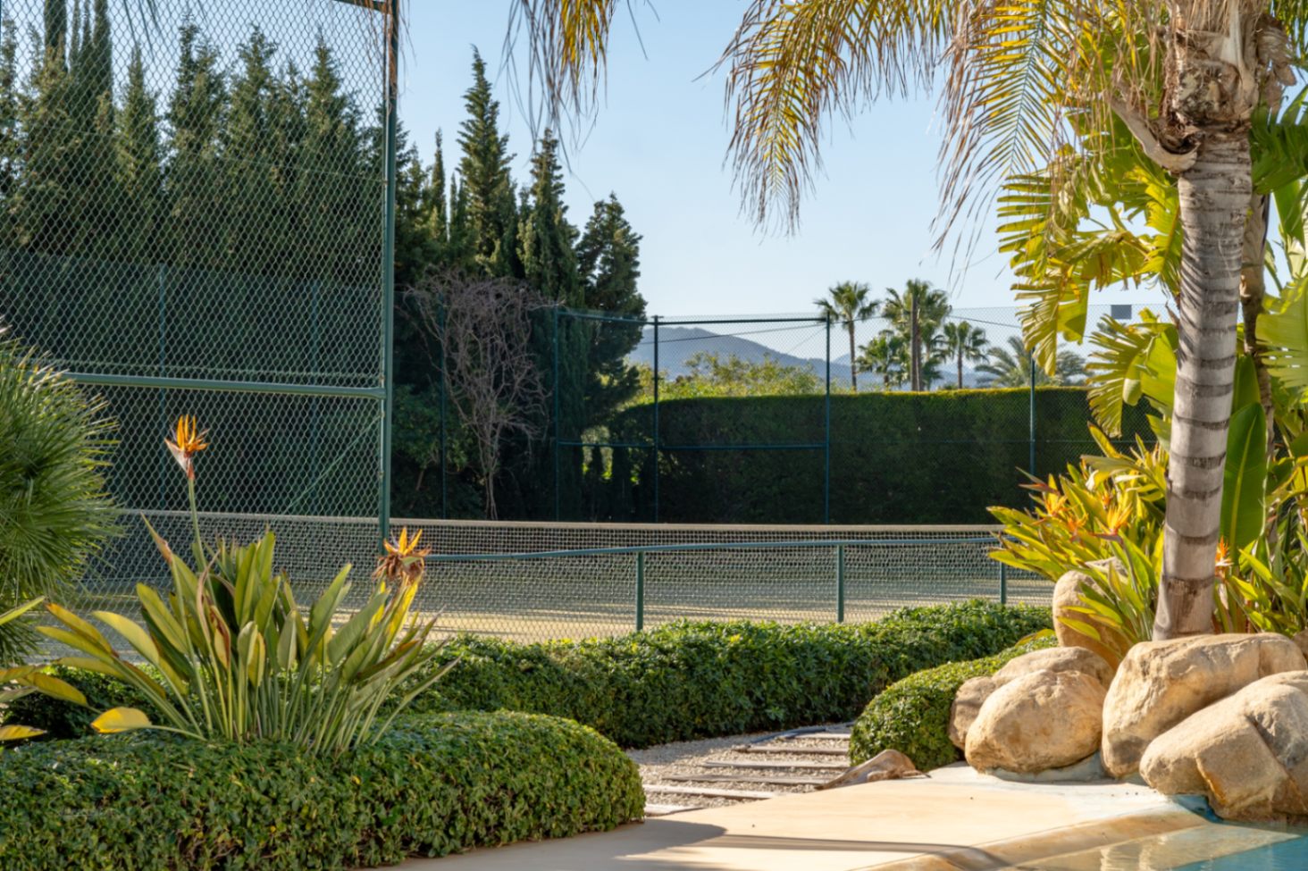 Court de tennis privé entouré de haies denses et d'une végétation méditerranéenne, offrant une vue sur le Montgó depuis les jardins paysagers.