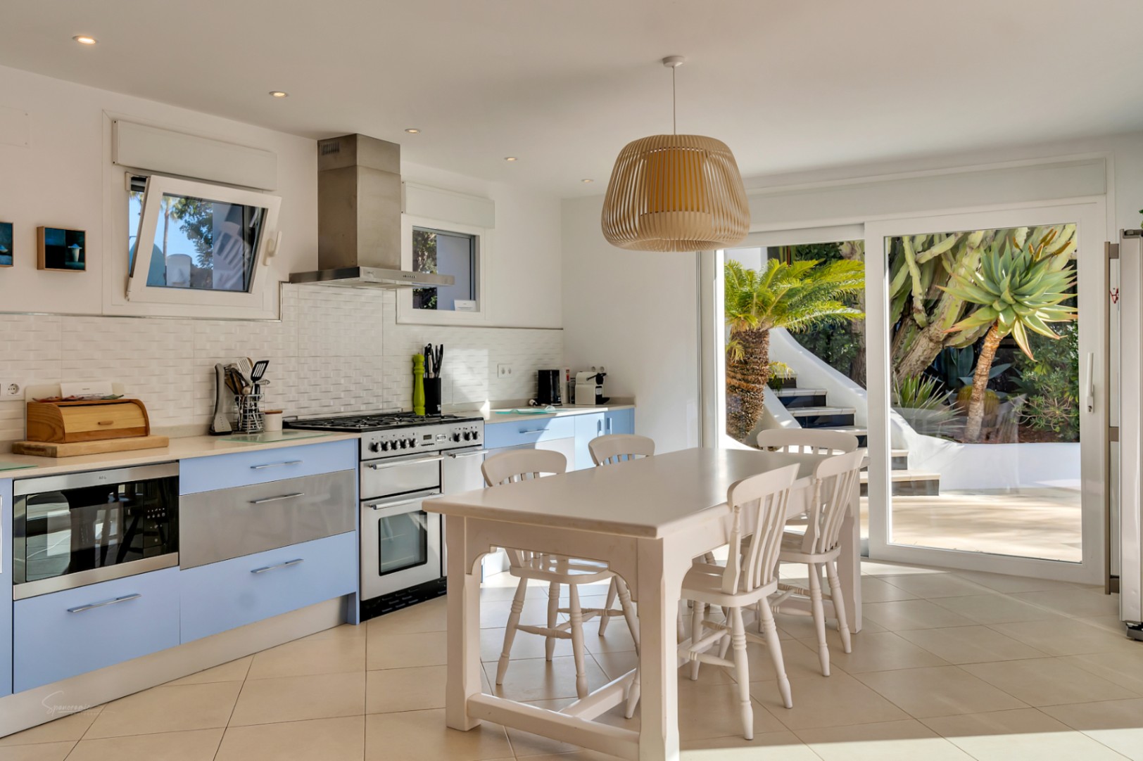 Cuisine méditerranéenne avec des meubles bleu clair, une cuisinière en inox et un coin repas. Les grandes baies vitrées offrent une vue directe sur la terrasse et le jardin.