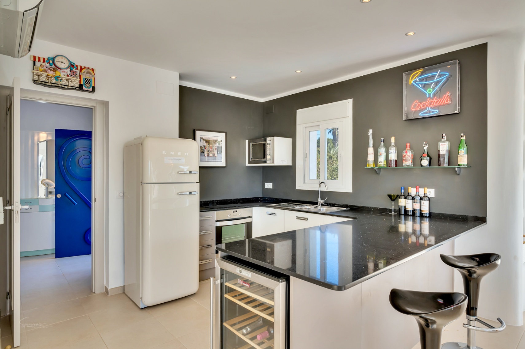 Cuisine moderne avec plans de travail en granit noir, réfrigérateur Smeg rétro et coin bar équipé d'une cave à vin intégrée et d'un néon décoratif.