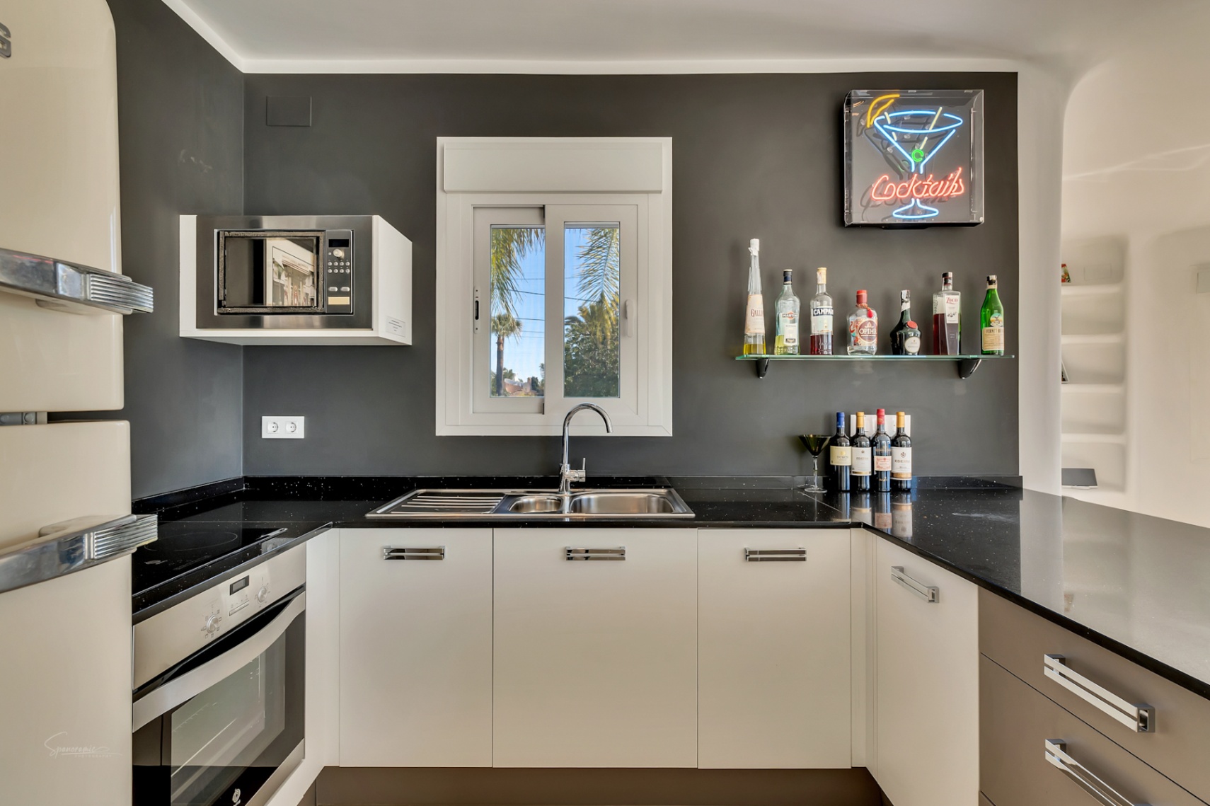 Cuisine moderne avec plans de travail noirs, armoires blanches et coin bar avec enseigne au néon. Équipée d'appareils intégrés et d'une fenêtre sur le jardin.