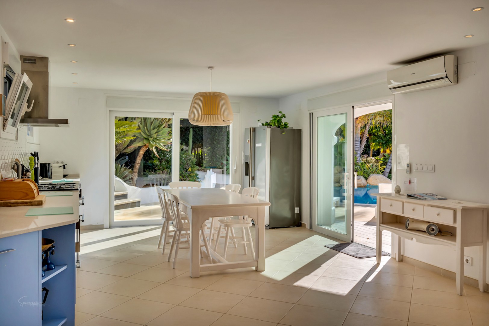 Cuisine ouverte et salle à manger avec table blanche et meubles bleus, offrant un accès direct à la piscine et au jardin via de larges baies vitrées coulissantes.
