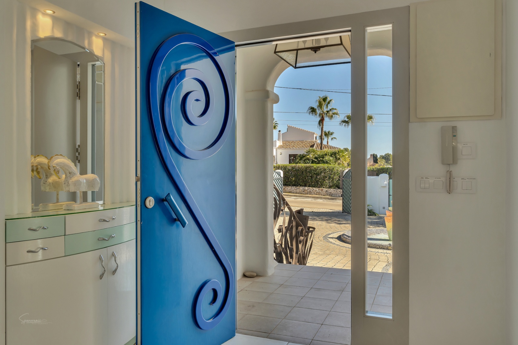 Entrée principale avec une porte bleue au design en spirale, s'ouvrant sur une cour pavée et un jardin méditerranéen ensoleillé.