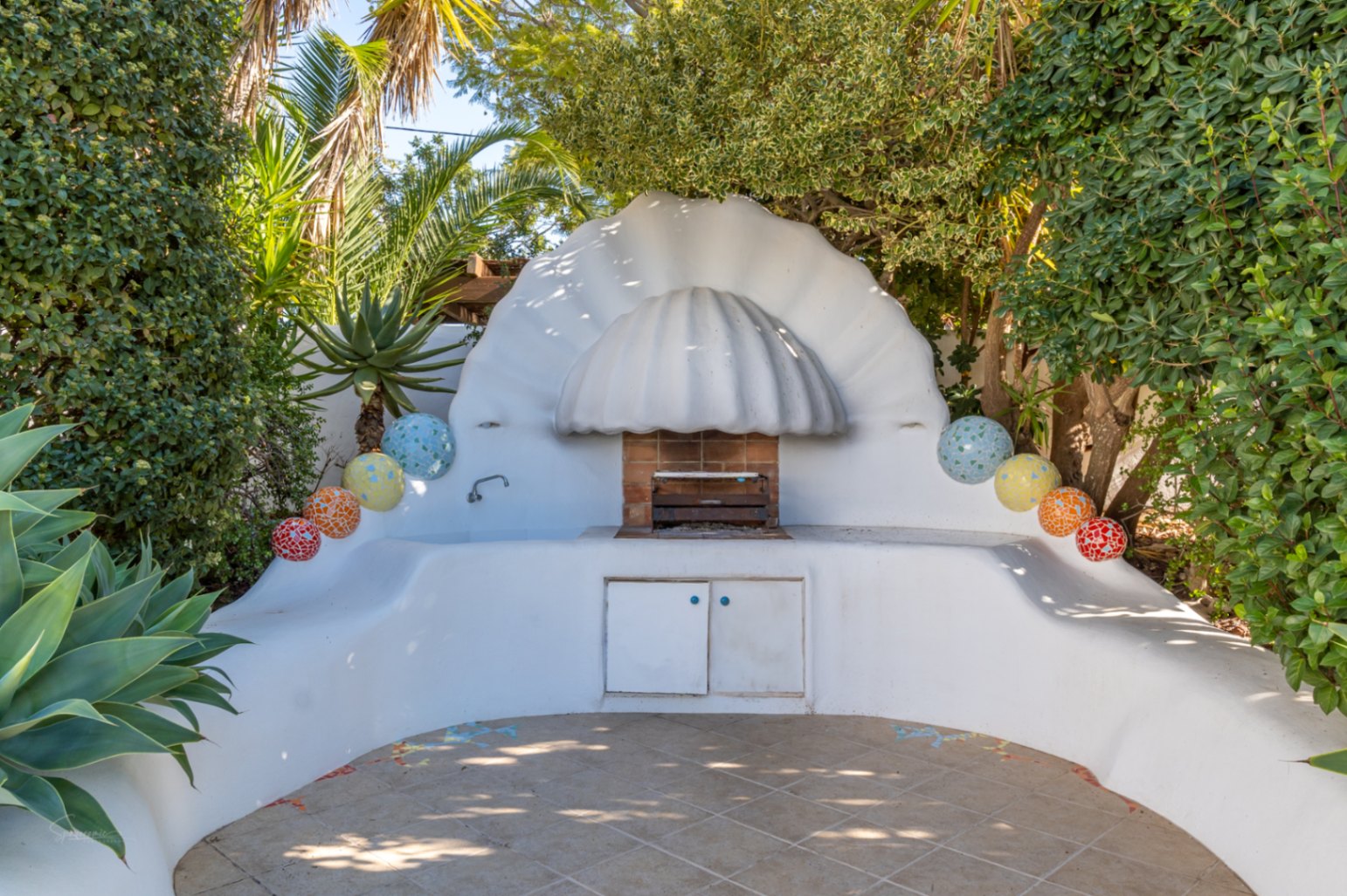 Espace barbecue extérieur maçonné avec une hotte sculpturale en forme de coquillage, agrémenté de sphères en mosaïque et entouré d'une végétation tropicale dense.