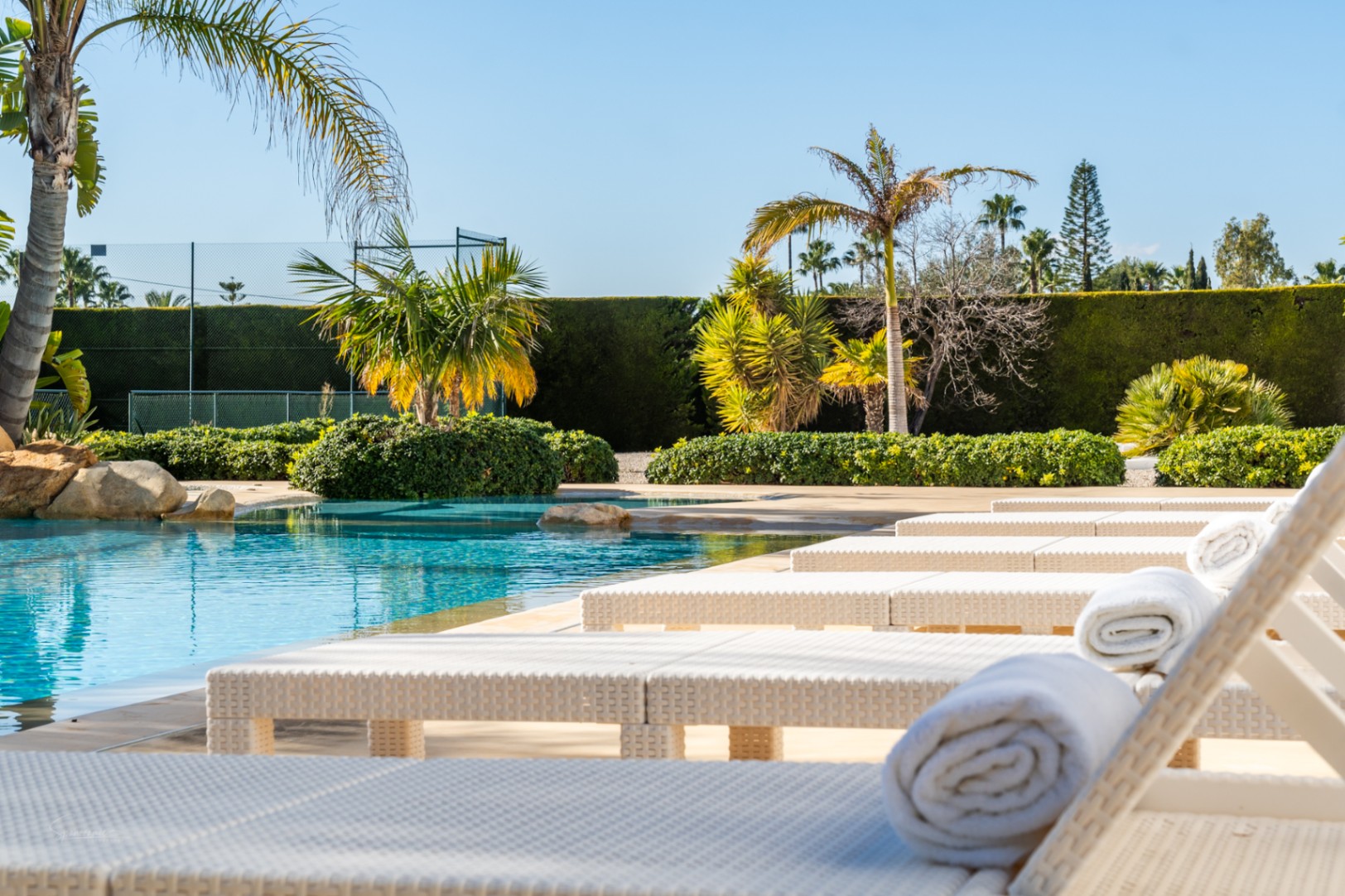 Espace de détente au bord de la piscine avec des bains de soleil en rotin blanc, entouré de palmiers tropicaux et d'un court de tennis privé.