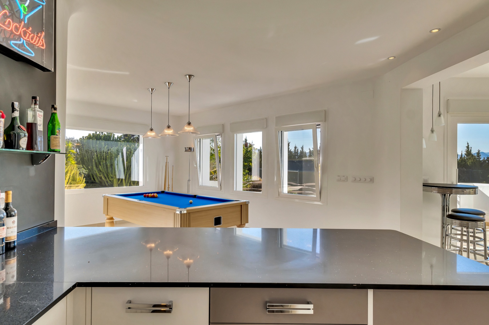Espace de loisirs avec billard au tapis bleu, comptoir de bar moderne et grandes fenêtres laissant entrer une lumière naturelle généreuse.