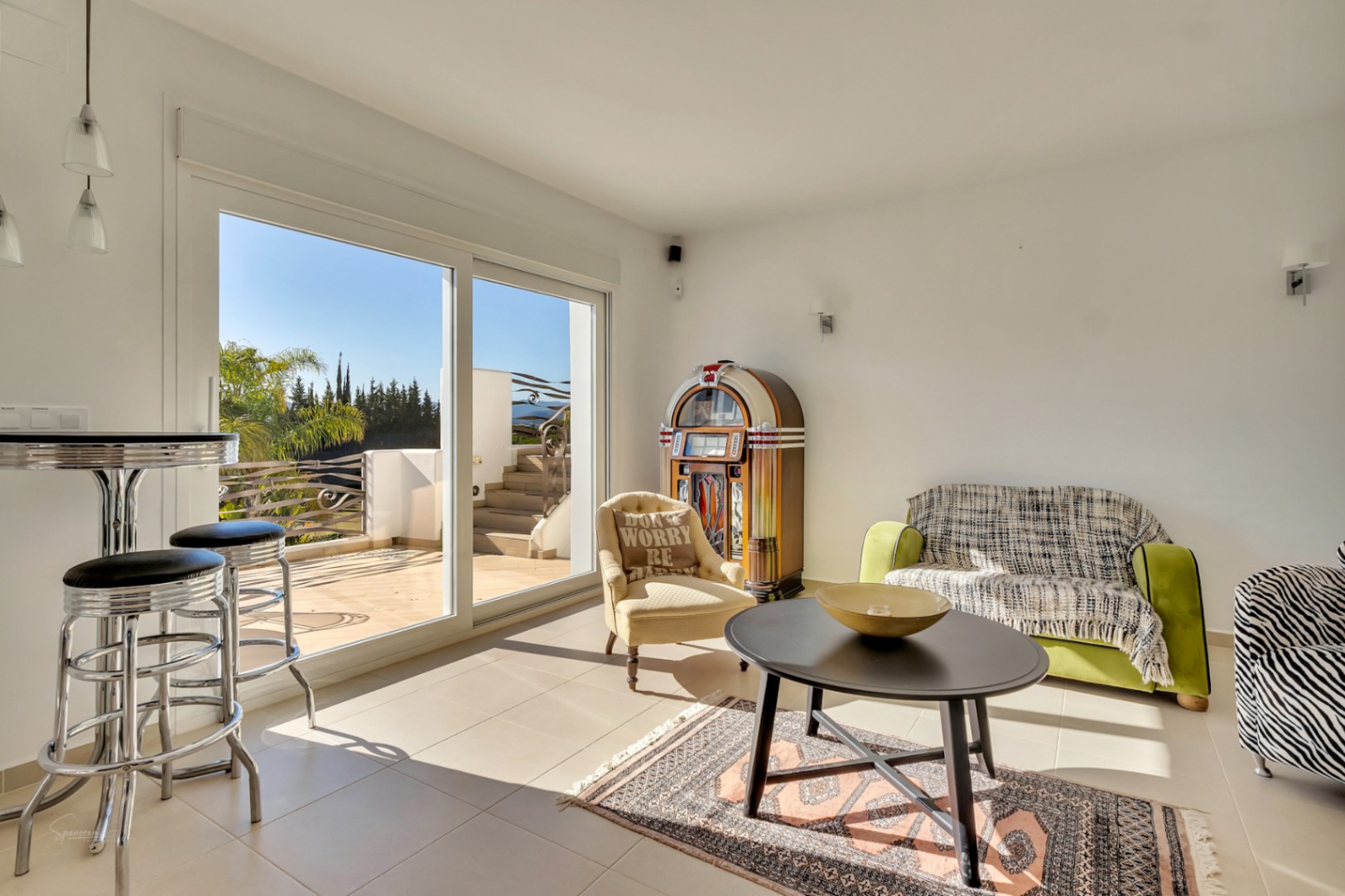 Espace de vie lumineux avec jukebox rétro, mobilier varié et grandes baies vitrées offrant un accès direct à une terrasse ensoleillée et dégagée.