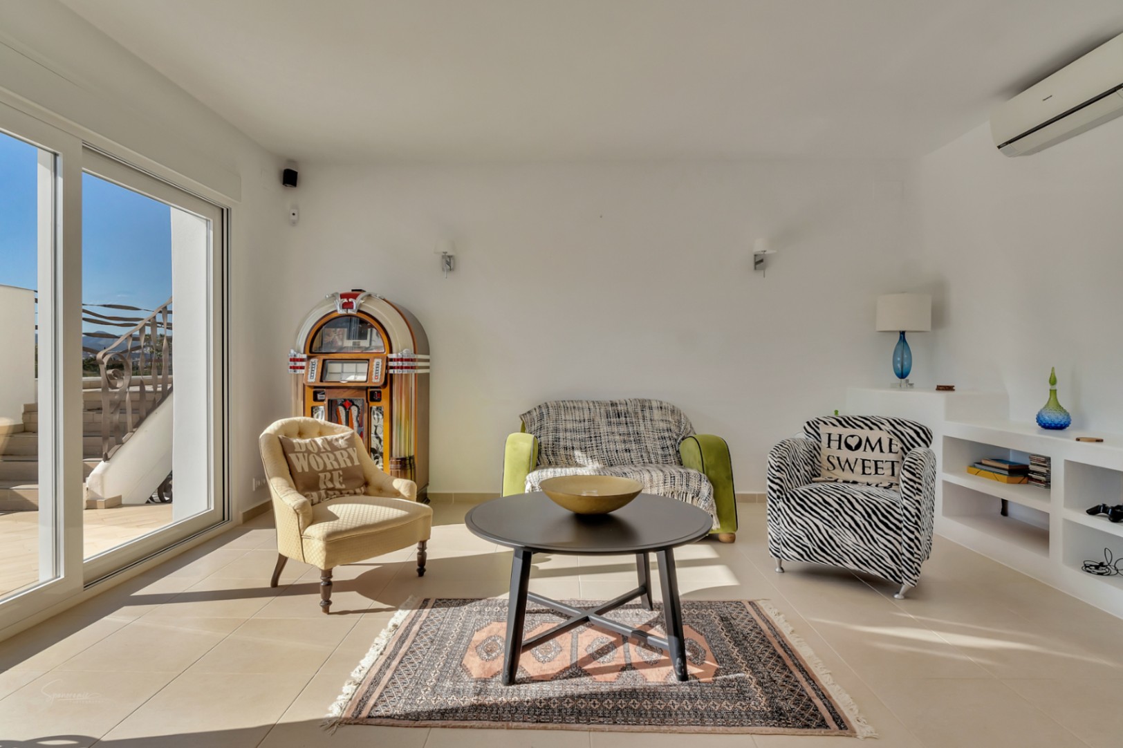 Espace de vie lumineux avec mobilier éclectique, jukebox vintage et étagères intégrées, offrant un accès direct à la terrasse par de grandes baies vitrées.
