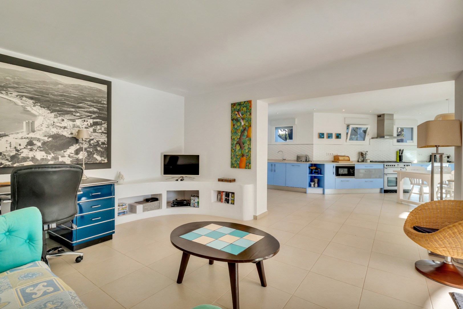 Espace de vie ouvert intégrant un coin bureau avec mobilier bleu et une cuisine moderne aux façades bleu clair, le tout sur un sol en carrelage uniforme.