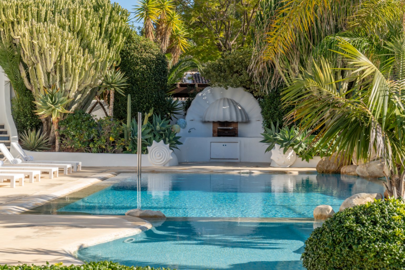 Espace piscine avec barbecue blanc au design organique, entouré d'une végétation méditerranéenne luxuriante, de grands cactus et de bains de soleil blancs.