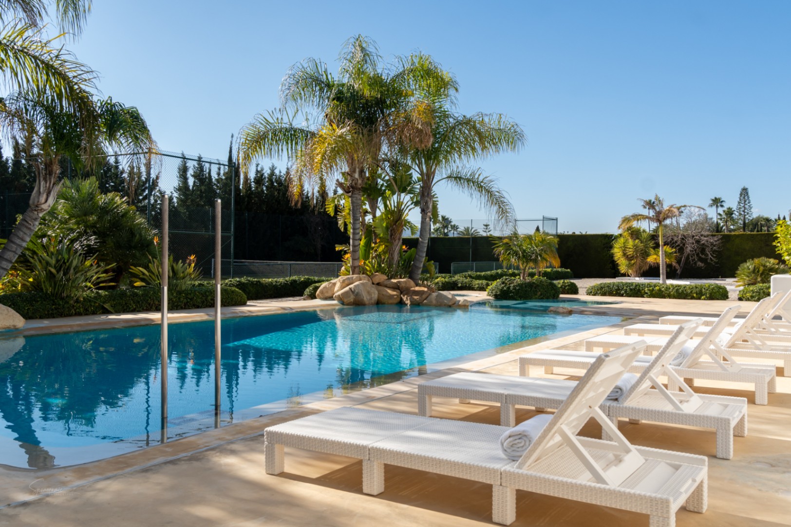 Espace piscine de 70m² avec bains de soleil blancs, palmiers tropicaux et enrochements naturels, offrant une vue sur le court de tennis privé et le jardin paysager.