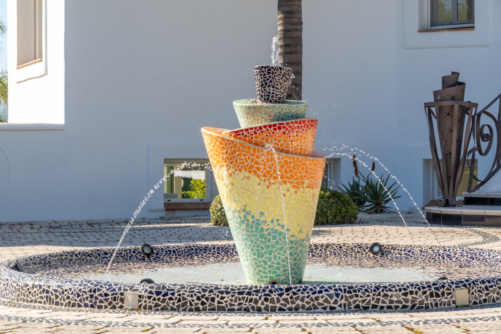 Fontaine centrale en mosaïque multicolore au design en spirale, située dans la cour d'entrée pavée devant la façade blanche épurée.