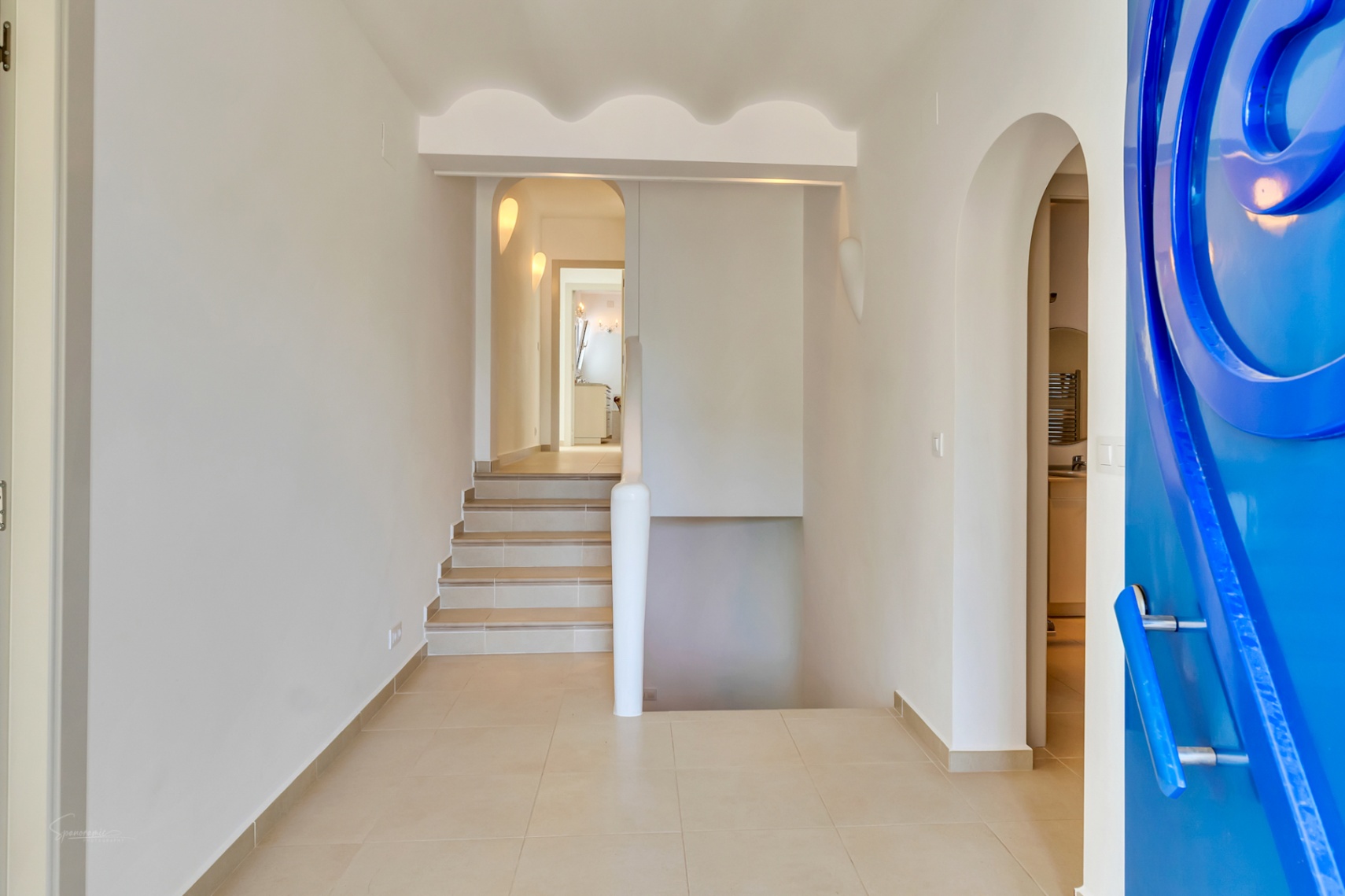 Hall d'entrée avec une porte bleue sculptée, un sol en grès cérame et un escalier menant à l'étage sous un plafond aux finitions ondulées élégantes.