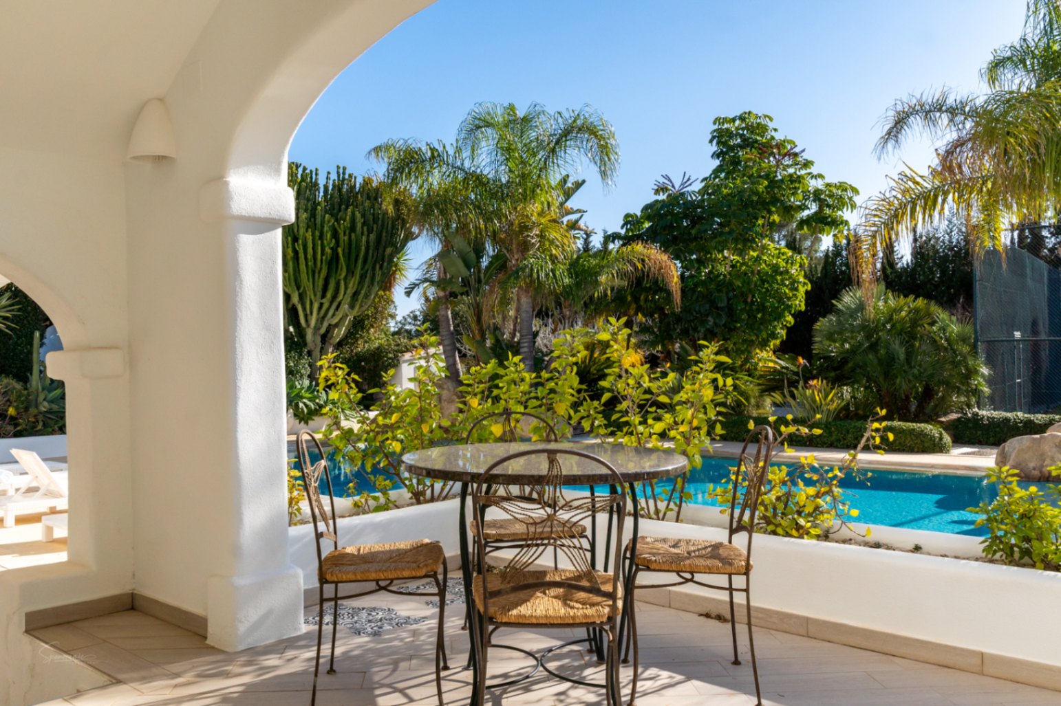 Naya de style Ibiza avec arches blanches et mobilier en fer forgé, offrant un espace d'ombre avec vue sur la piscine et le jardin tropical luxuriant.