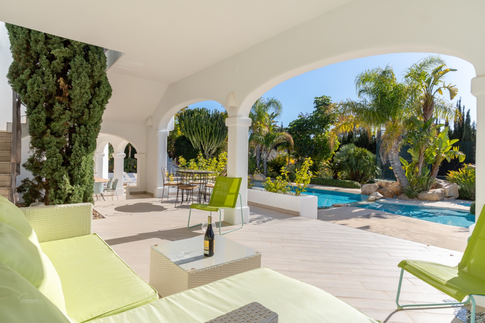 Naya ombragée aux arches blanches avec mobilier vert citron, offrant une vue sur la piscine et le jardin méditerranéen arboré de palmiers.