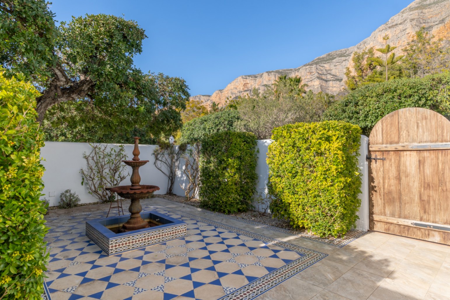 Patio de style andalou avec carrelage géométrique bleu et blanc, fontaine en fer à étages et vue sur le mont Montgó derrière les murs blancs.