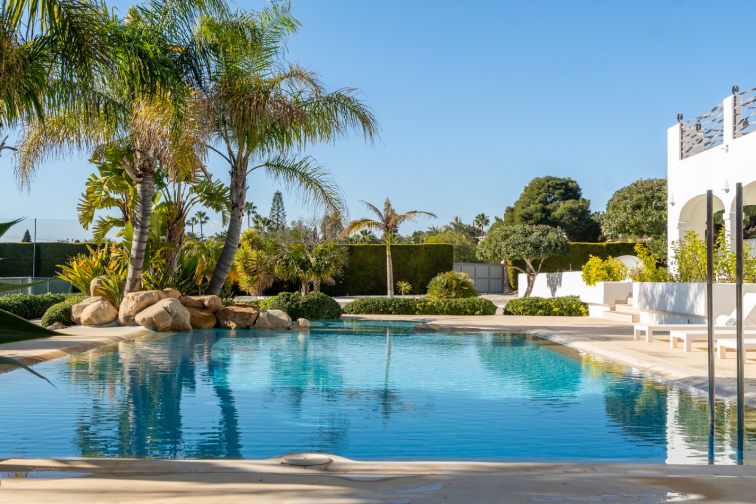 Piscine paysagère avec entrée progressive, bordée de palmiers et de rochers, offrant une vue sur la naya aux arches blanches et le court de tennis privé.