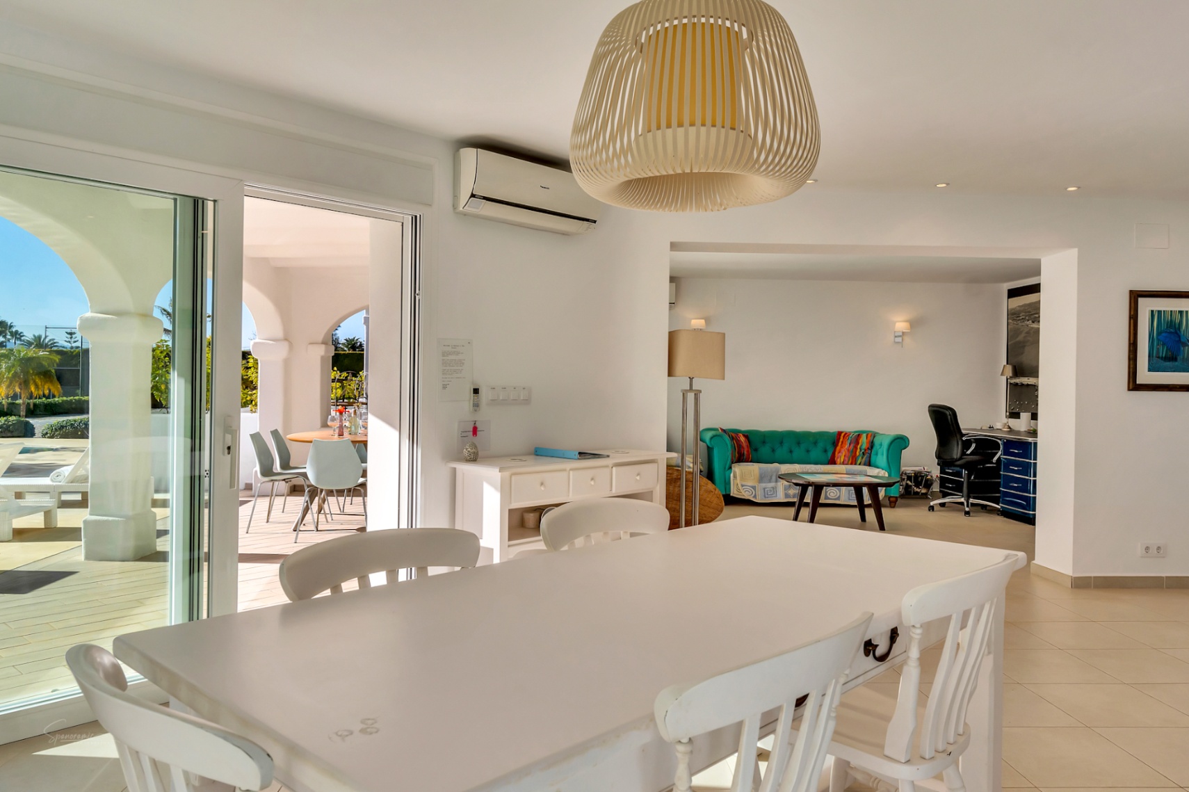 Salle à manger lumineuse avec table en bois blanc et suspension en osier, ouverte sur un salon avec canapé turquoise, coin bureau et accès à la terrasse.