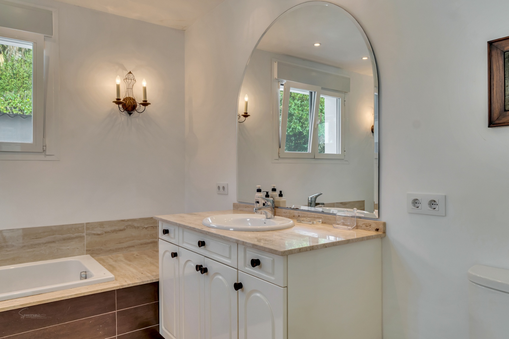 Salle de bain avec plan de travail en marbre, grand miroir en arche et appliques murales. Elle dispose d'une baignoire intégrée et d'une fenêtre offrant une lumière naturelle.