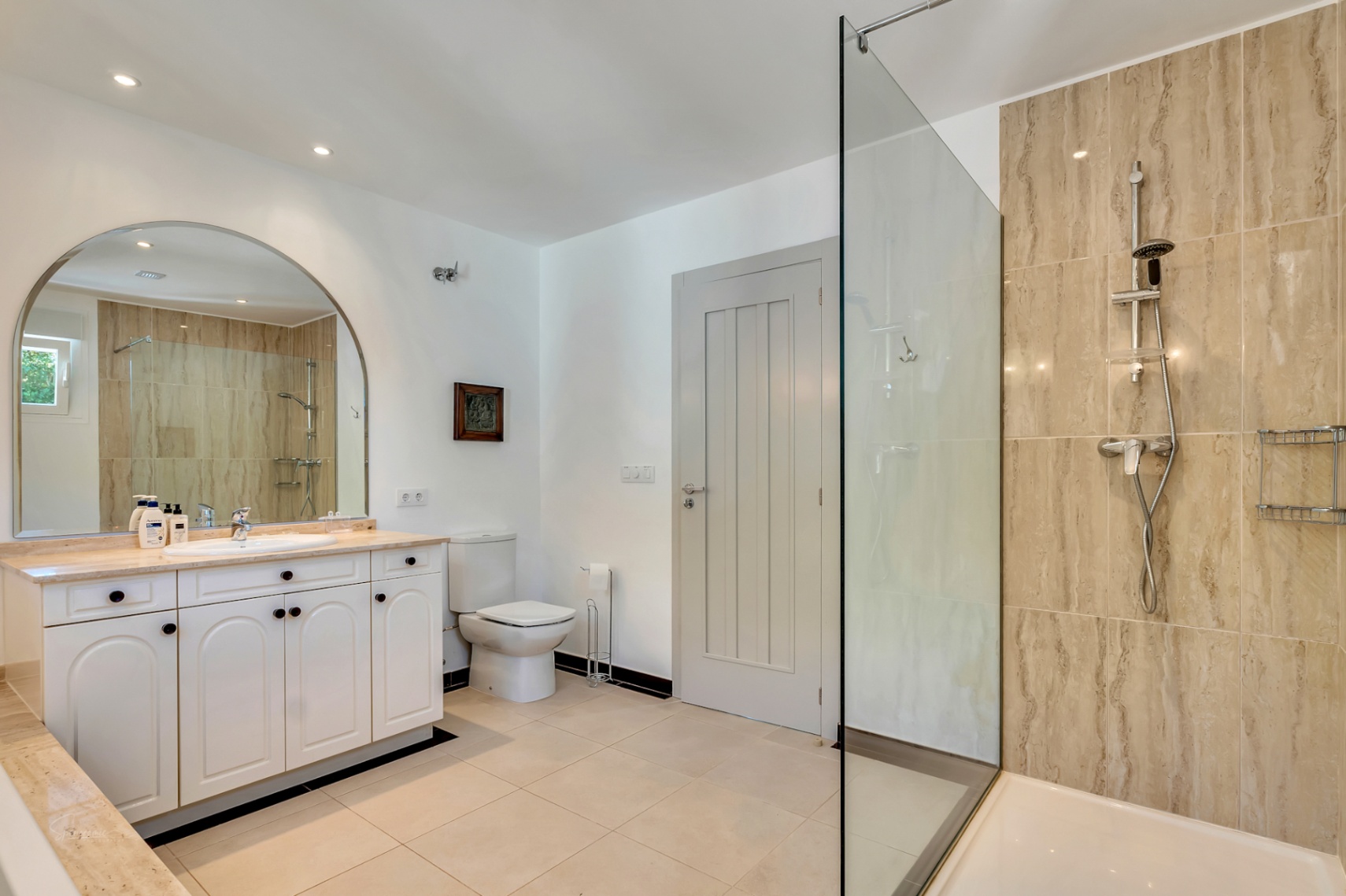 Salle de bain élégante avec douche à l'italienne en pierre naturelle, meuble vasque blanc avec plan de travail en marbre et grand miroir cintré.
