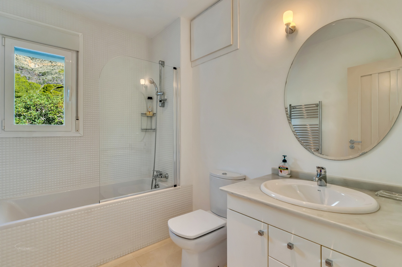 Salle de bain lumineuse avec mosaïque blanche, baignoire avec paroi vitrée et plan en marbre. La fenêtre offre une vue dégagée sur la montagne Montgó.