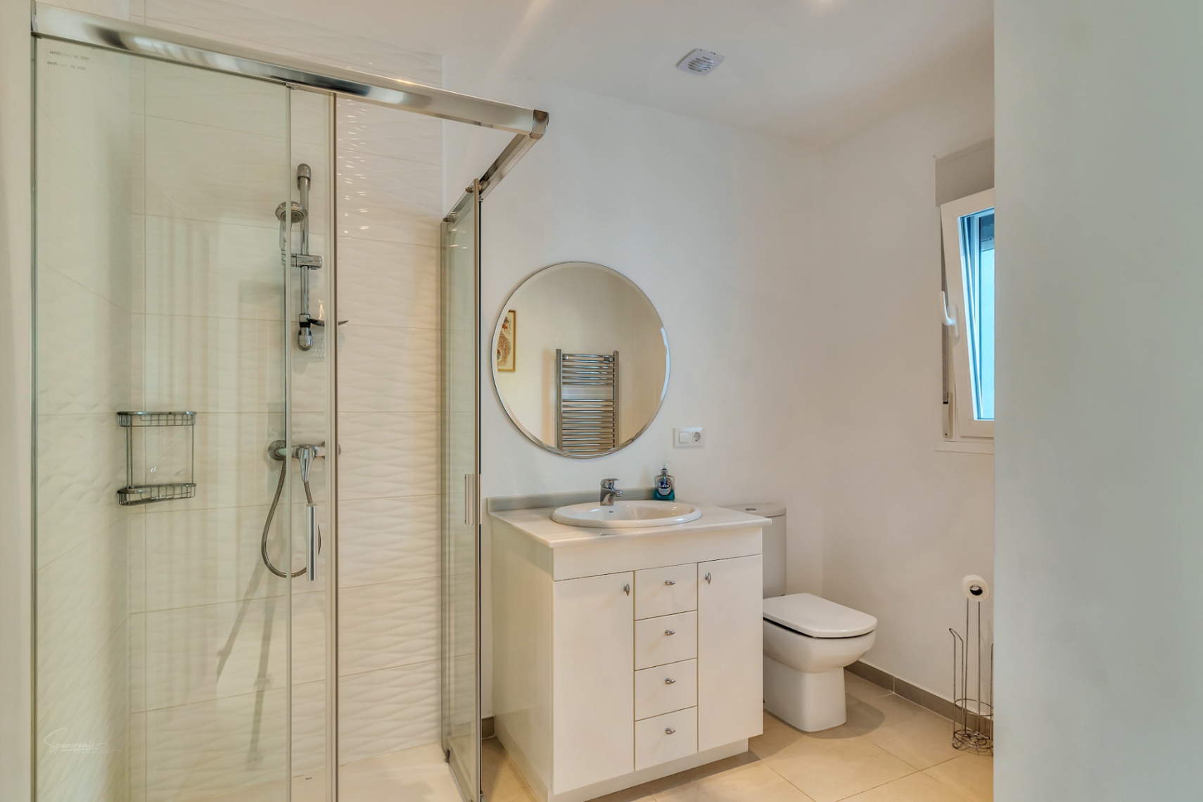 Salle de bain moderne avec douche vitrée, carrelage texturé, meuble vasque blanc surmonté d'un miroir circulaire et fenêtre latérale.