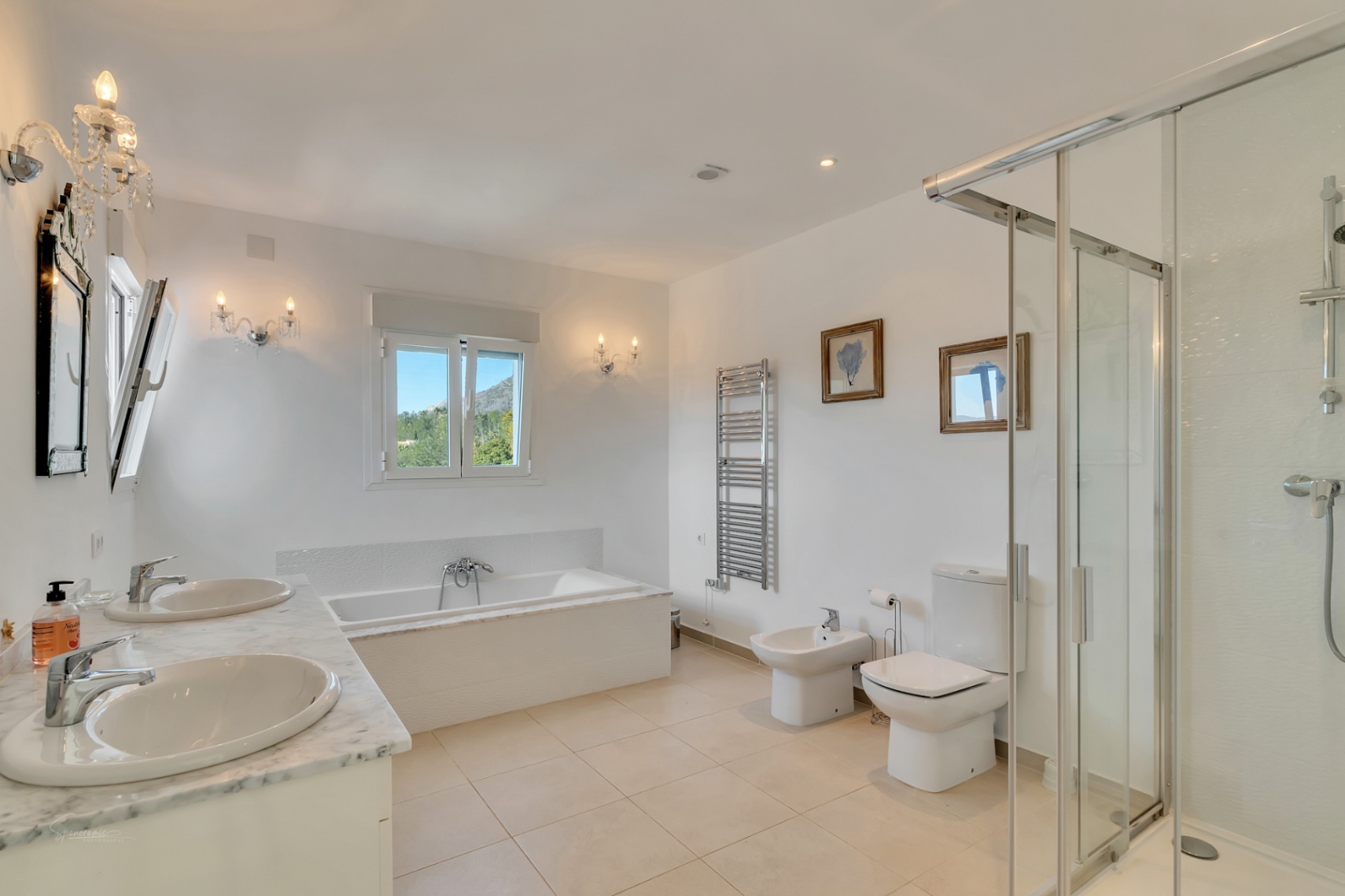 Salle de bain principale avec double vasque en marbre, baignoire intégrée, douche vitrée et appliques en cristal offrant une vue sur le Montgó.