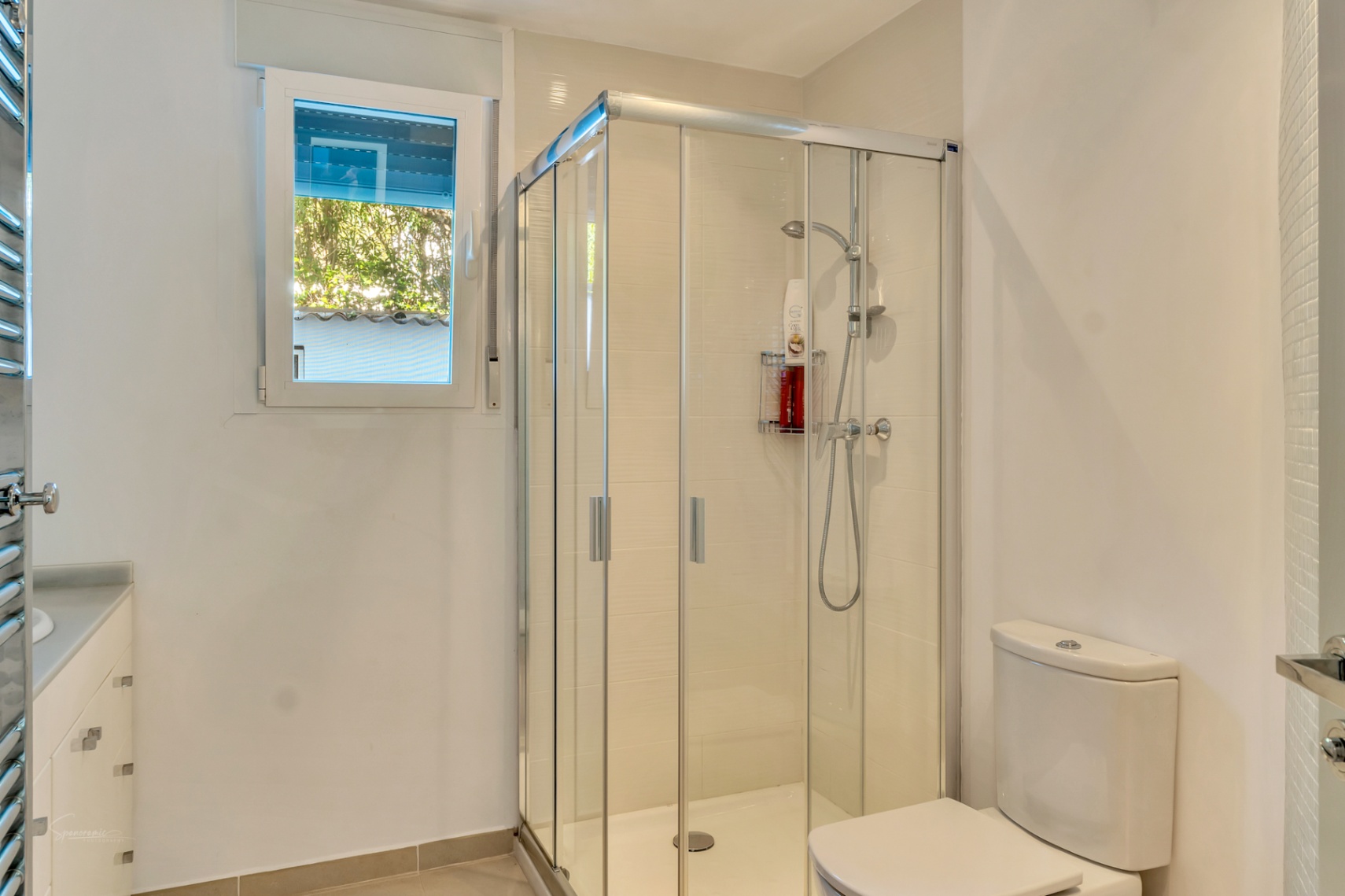 Salle de bains moderne avec cabine de douche en verre, sanitaires blancs et fenêtre offrant une lumière naturelle. Comprend un meuble vasque avec rangements et un sèche-serviettes.