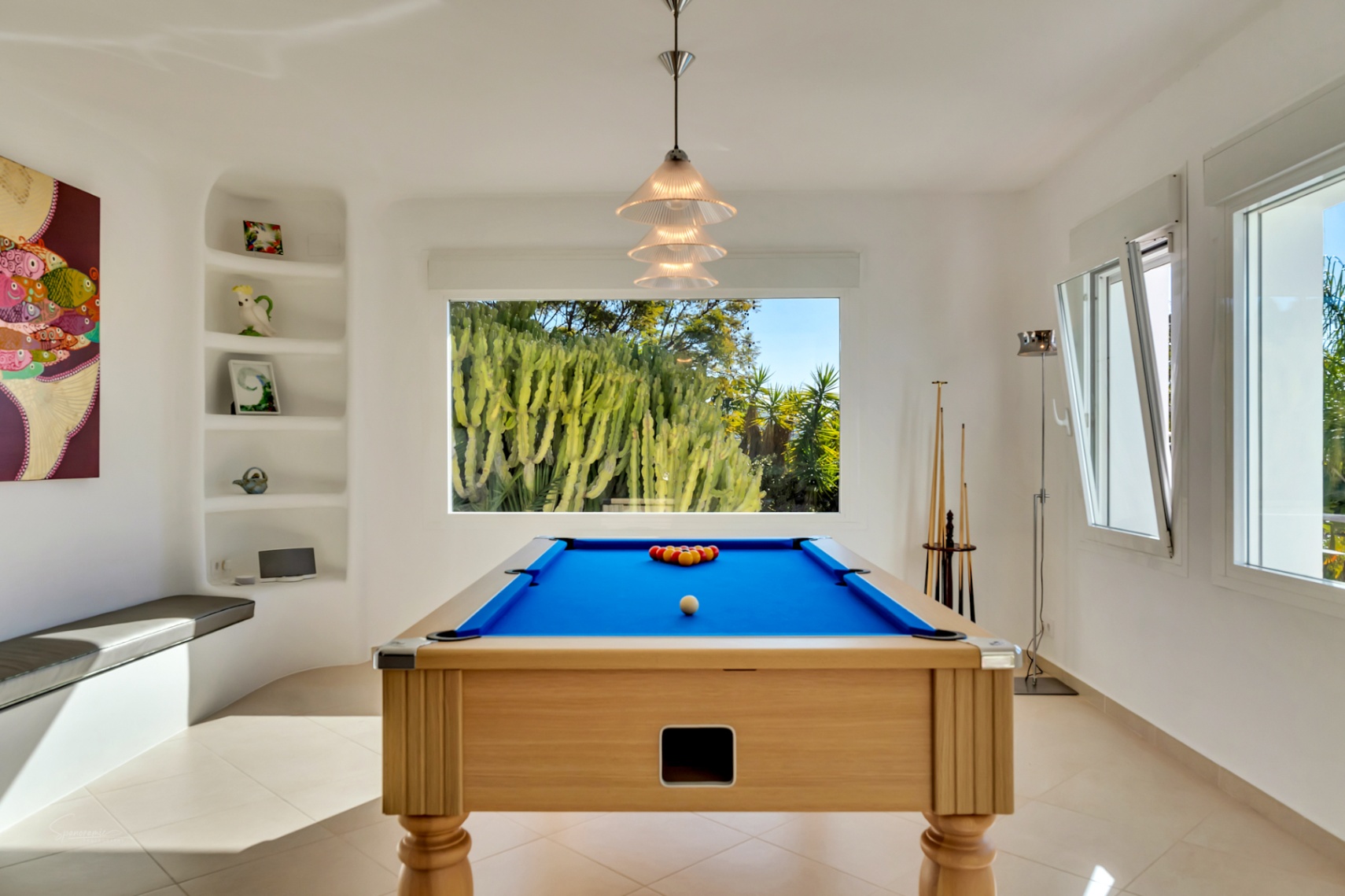 Salle de jeux avec billard en bois et tapis bleu, dotée d'étagères encastrées et d'une grande fenêtre offrant une vue sur le jardin de cactus.