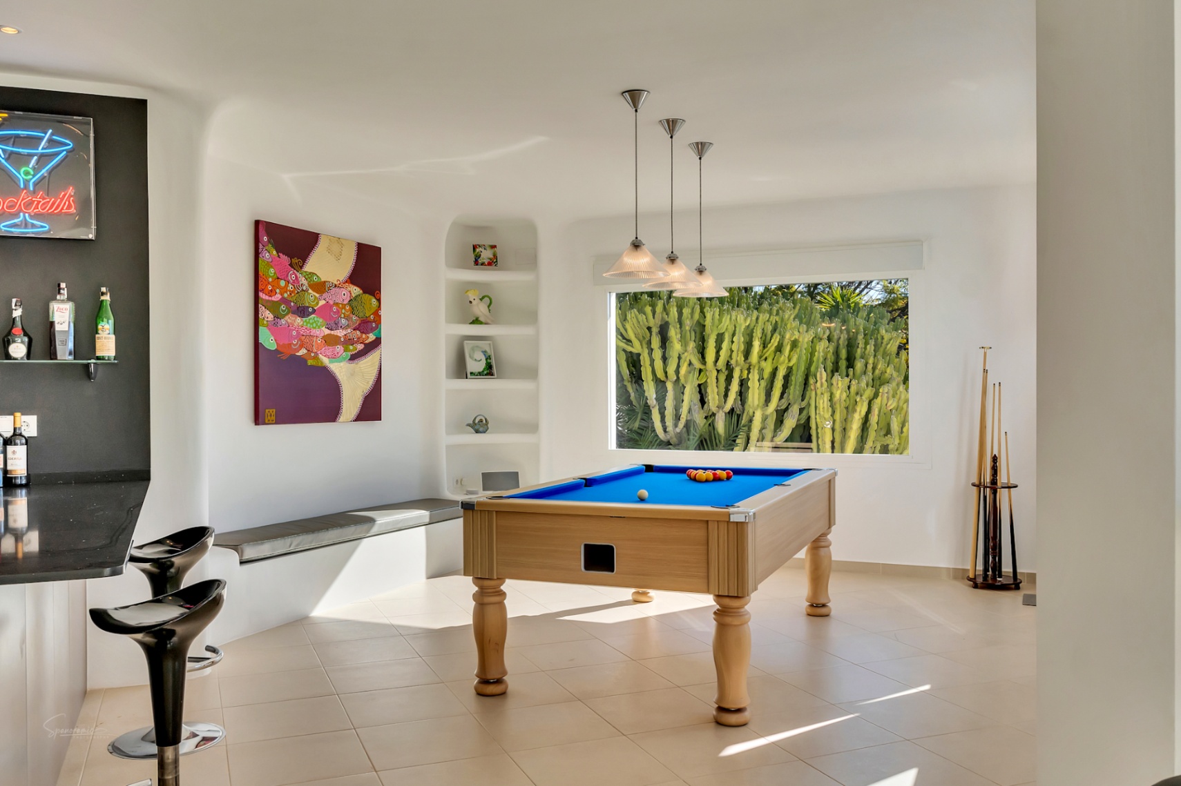 Salle de jeux comprenant un billard, un espace bar avec tabourets et une grande fenêtre offrant une vue sur les cactus. Murs blancs arrondis de style Ibiza et éclairage suspendu.