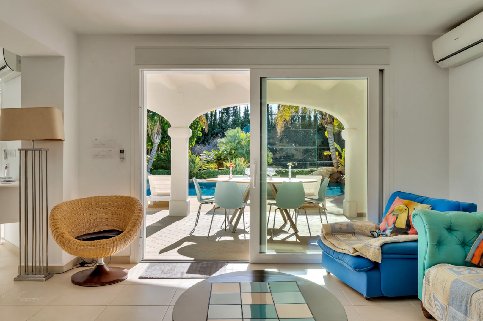 Salon lumineux s'ouvrant sur une naya couverte aux arches blanches, offrant une vue dégagée sur la piscine et le jardin méditerranéen verdoyant.