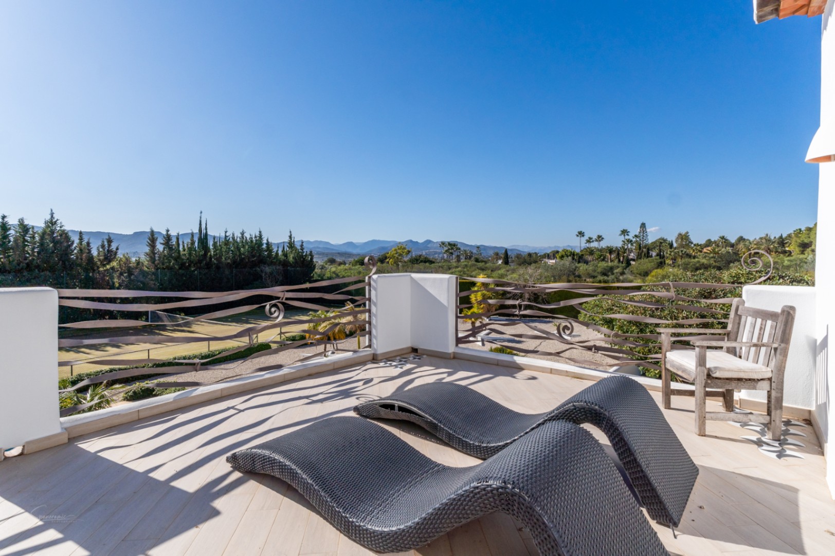 Terrasse supérieure avec bains de soleil ergonomiques et garde-corps en fer forgé, offrant une vue sur le court de tennis et le massif du Montgó.
