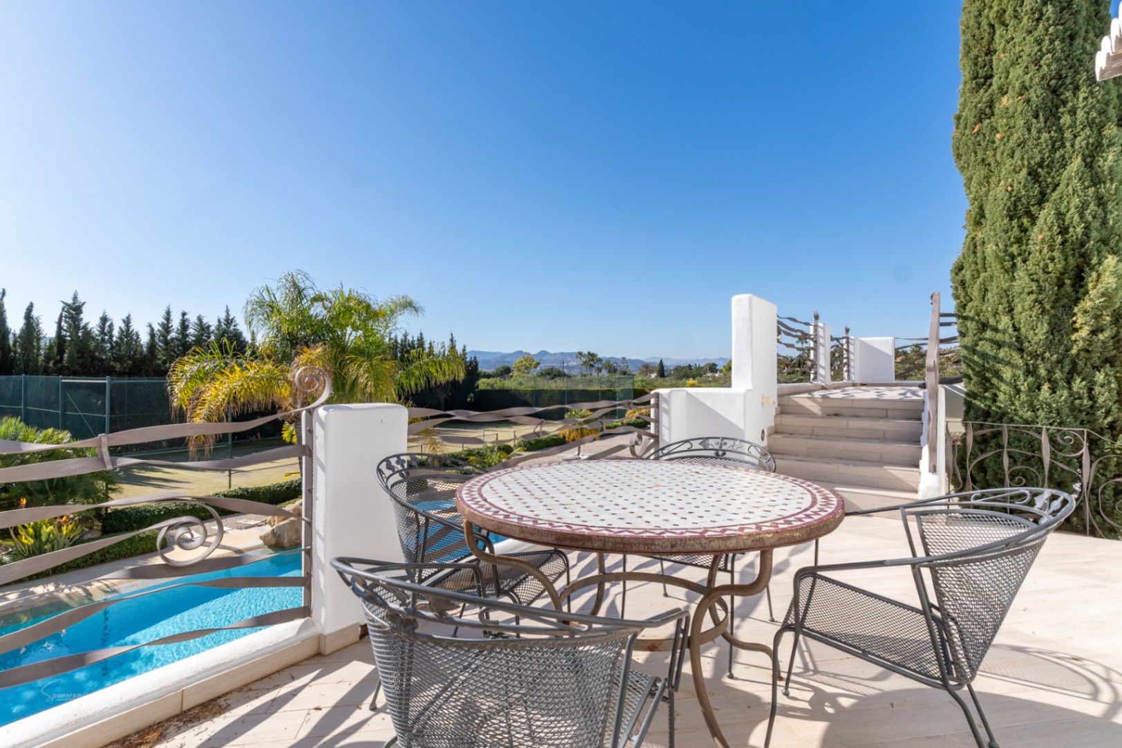 Terrasse supérieure avec table en mosaïque et fer forgé, offrant une vue dégagée sur la piscine, le court de tennis et la montagne Montgó.