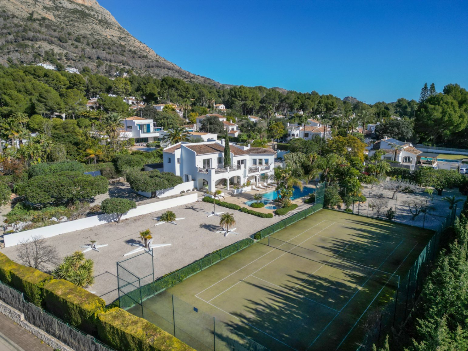 Vue aérienne de la villa de style Ibiza avec court de tennis privé, piscine bordée de palmiers et la montagne Montgó en arrière-plan.