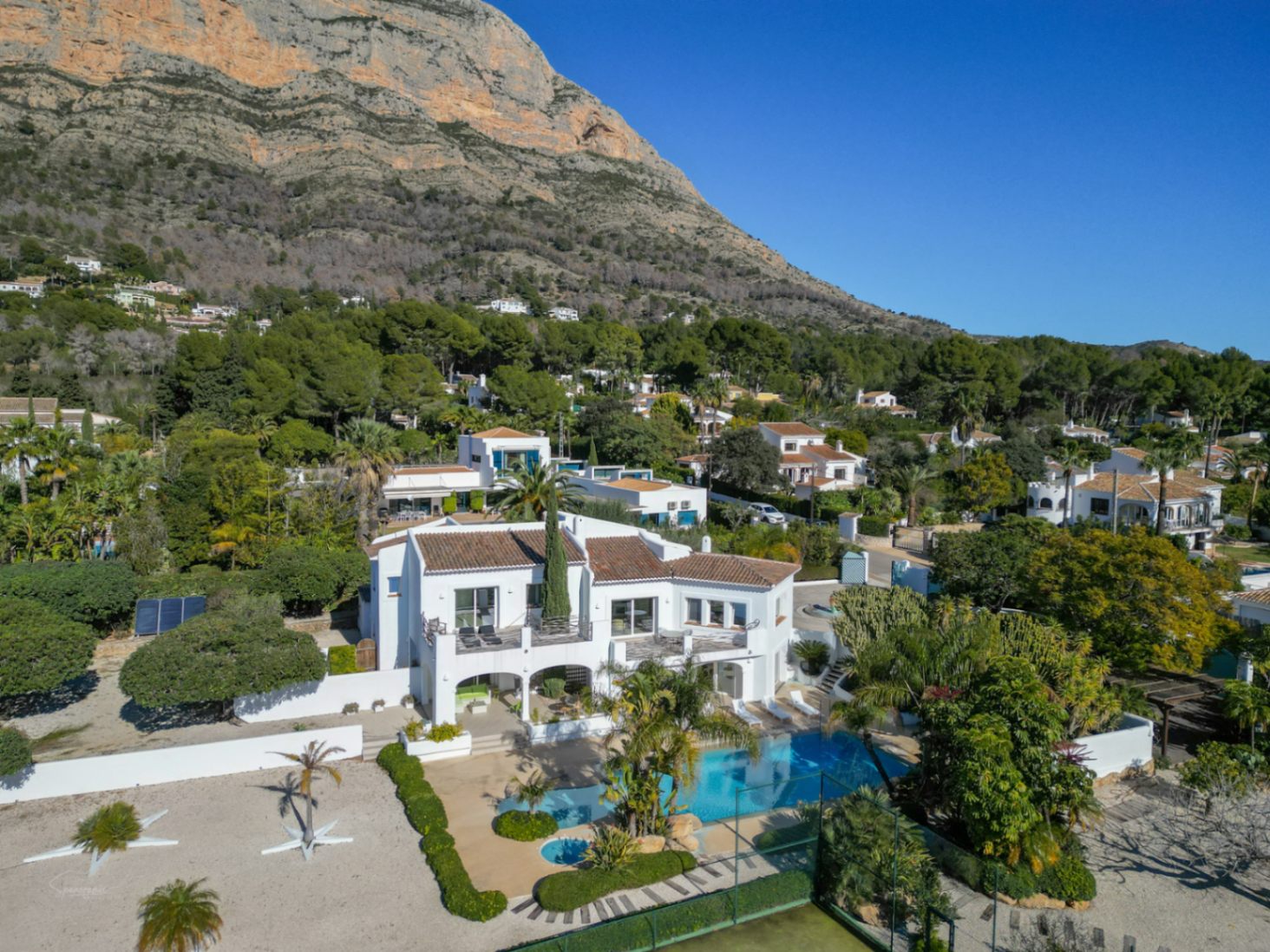 Vue aérienne de la villa de style Ibiza avec sa façade blanche, sa piscine, son court de tennis et le massif du Montgó en arrière-plan.
