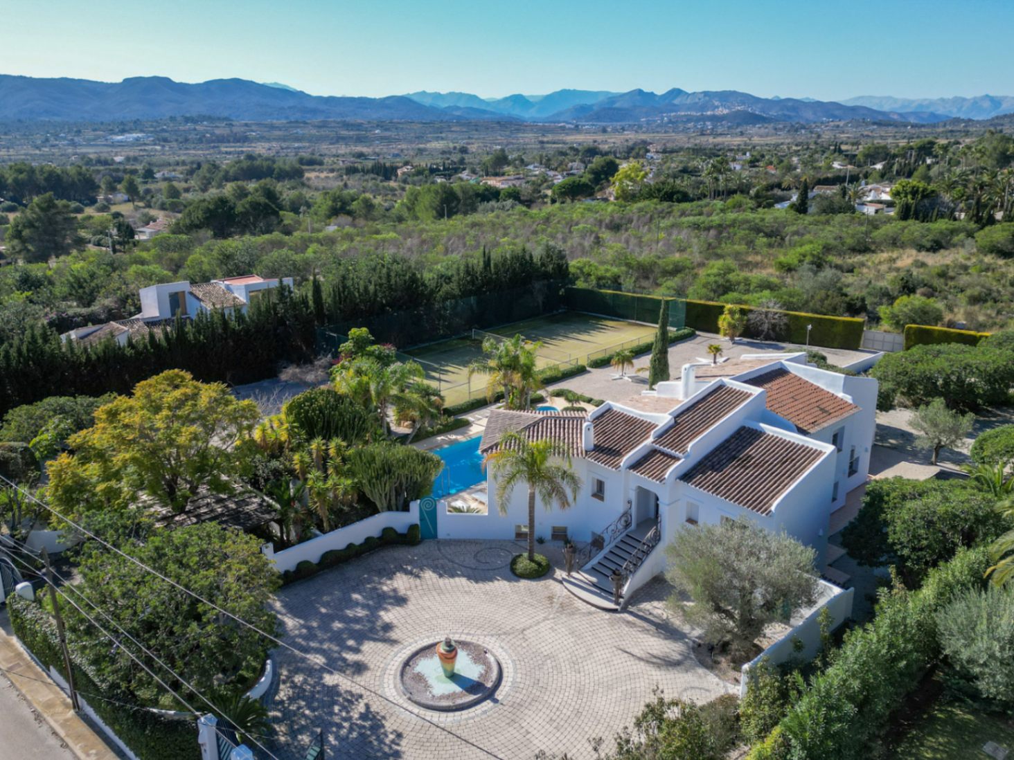 Vue aérienne de la villa de style Ibiza avec son entrée pavée circulaire, sa fontaine centrale, sa piscine, son court de tennis et ses vues sur la vallée du Montgó.