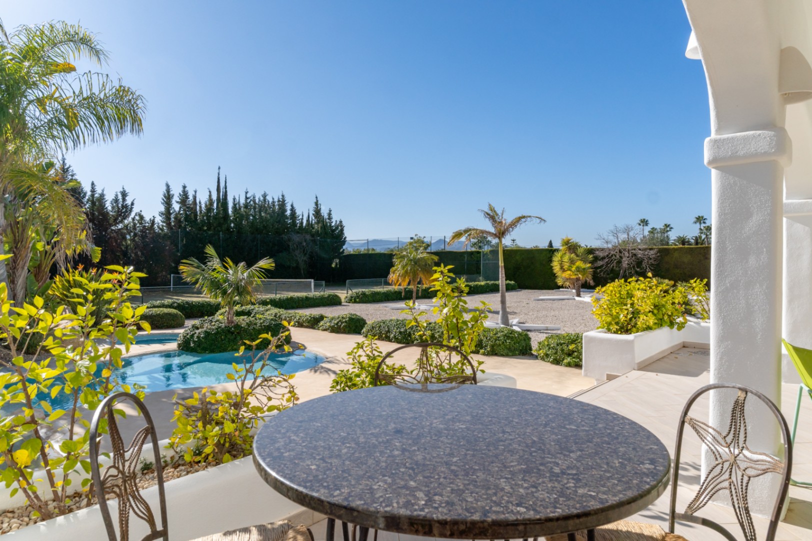 Vue depuis la naya avec table en granit, donnant sur la piscine, les jardins méditerranéens paysagers et le court de tennis privé.