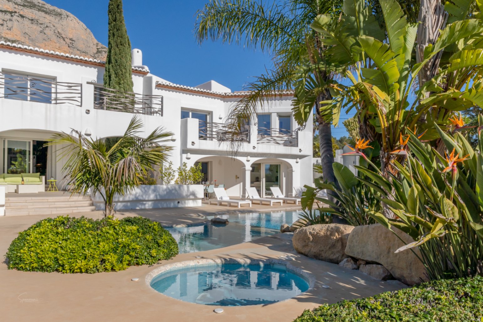 Vue extérieure de la villa de style Ibiza avec piscine, jacuzzi intégré et jardins tropicaux. La naya voûtée et les terrasses offrent une vue dégagée sur le mont Montgó.