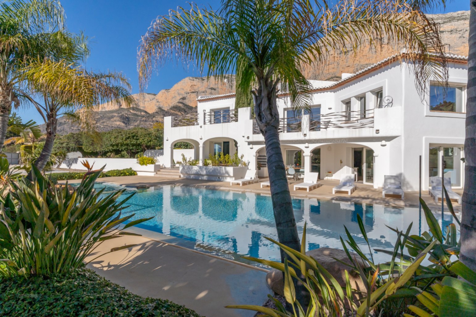 Vue extérieure de la villa de style Ibiza avec sa piscine, ses porches à arcades et ses terrasses à l'étage, sur fond de montagne Montgó et palmiers.