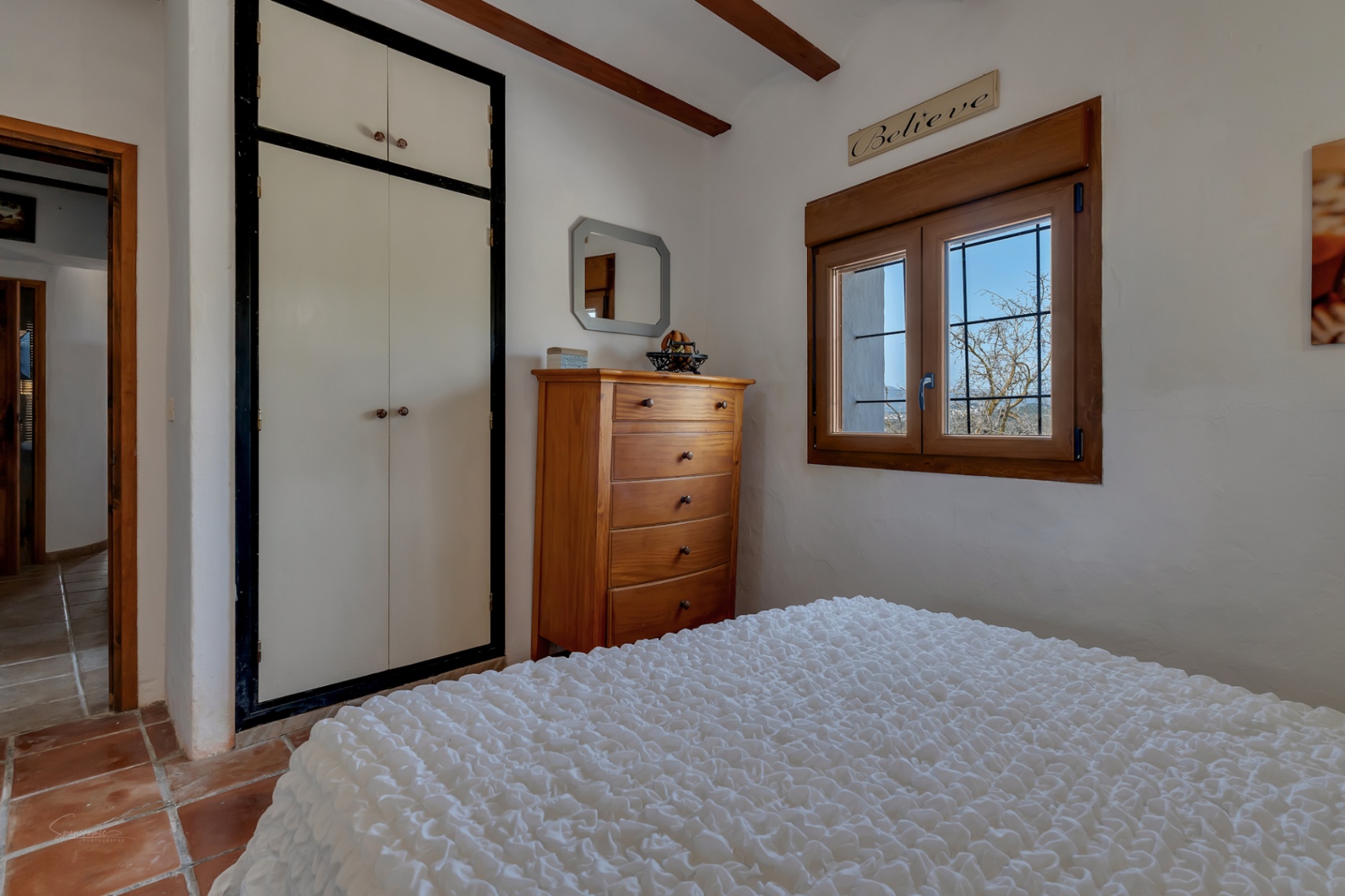 Chambre avec armoire encastrée blanche, commode en bois et fenêtre avec menuiseries bois.