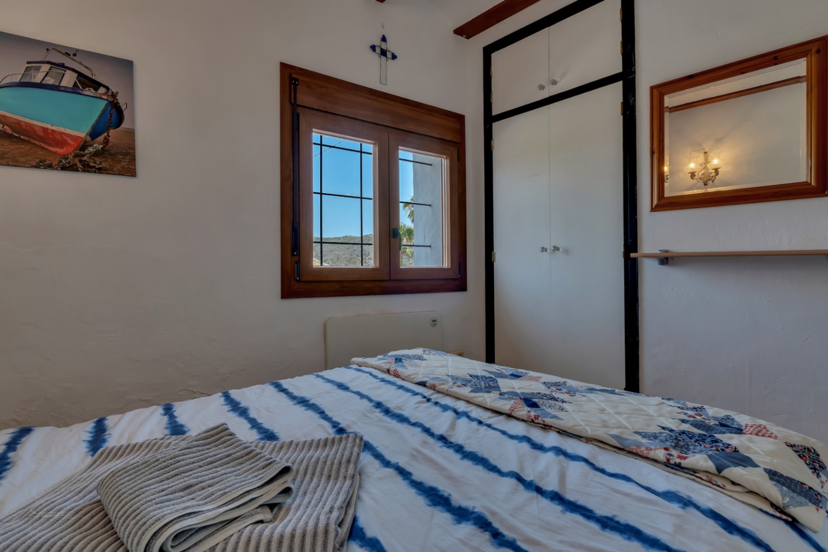 Chambre avec fenêtre en bois, placard blanc et literie à rayures bleues. Décor rustique et ambiance lumineuse.
