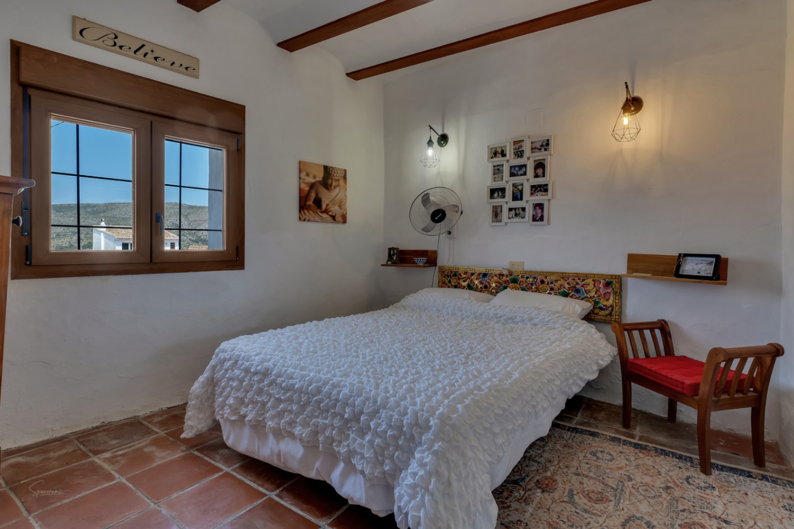 Chambre rustique avec sol en terre cuite, lit double avec tête de lit sculptée et fenêtre avec vue sur l'extérieur.