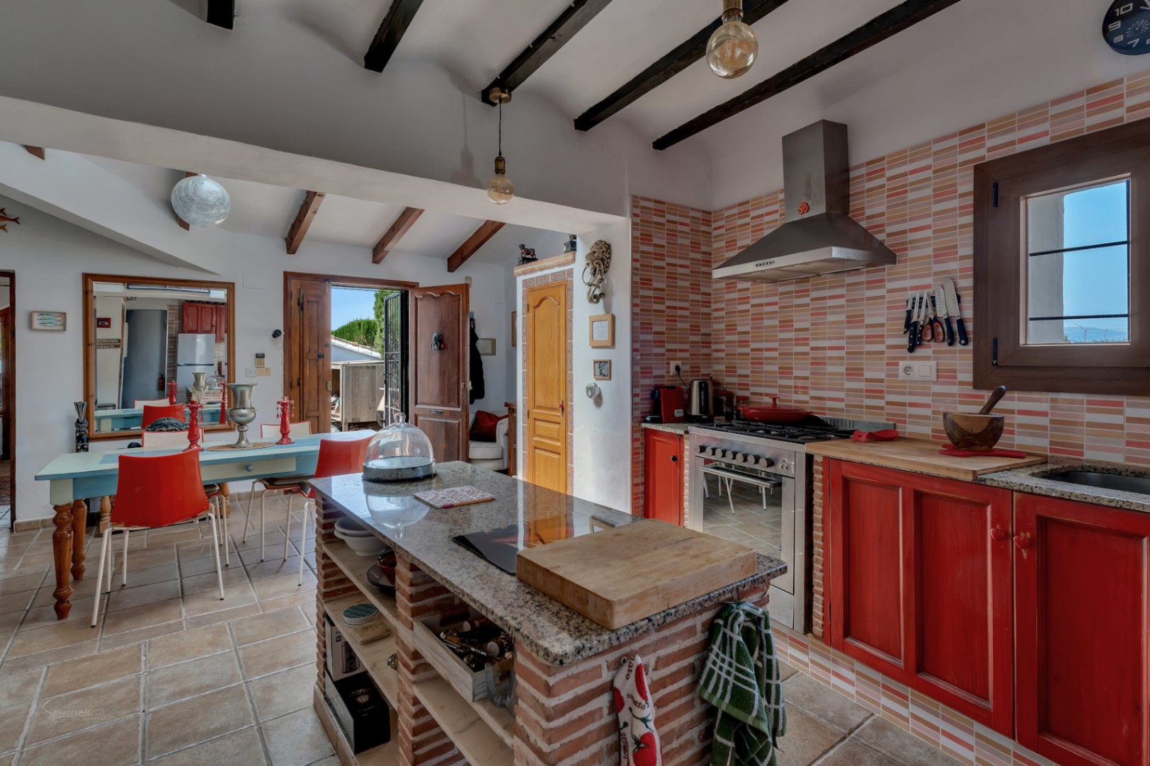 Cuisine rustique avec îlot en brique, crédence en carreaux pêche et meubles rouges. Poutres apparentes.