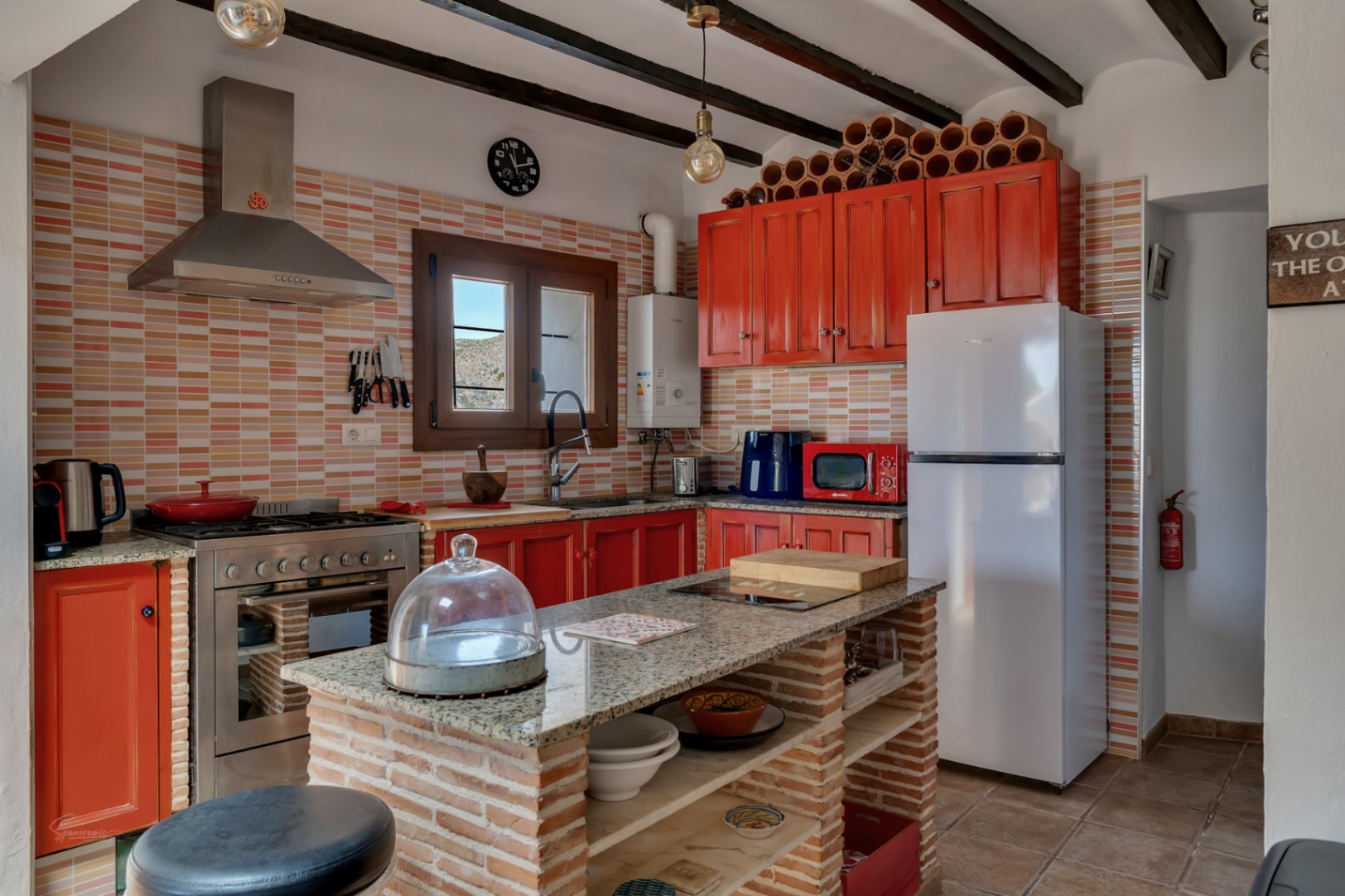 Cuisine rustique avec plans de travail en granit, armoires rouges et îlot central en brique avec étagères ouvertes.