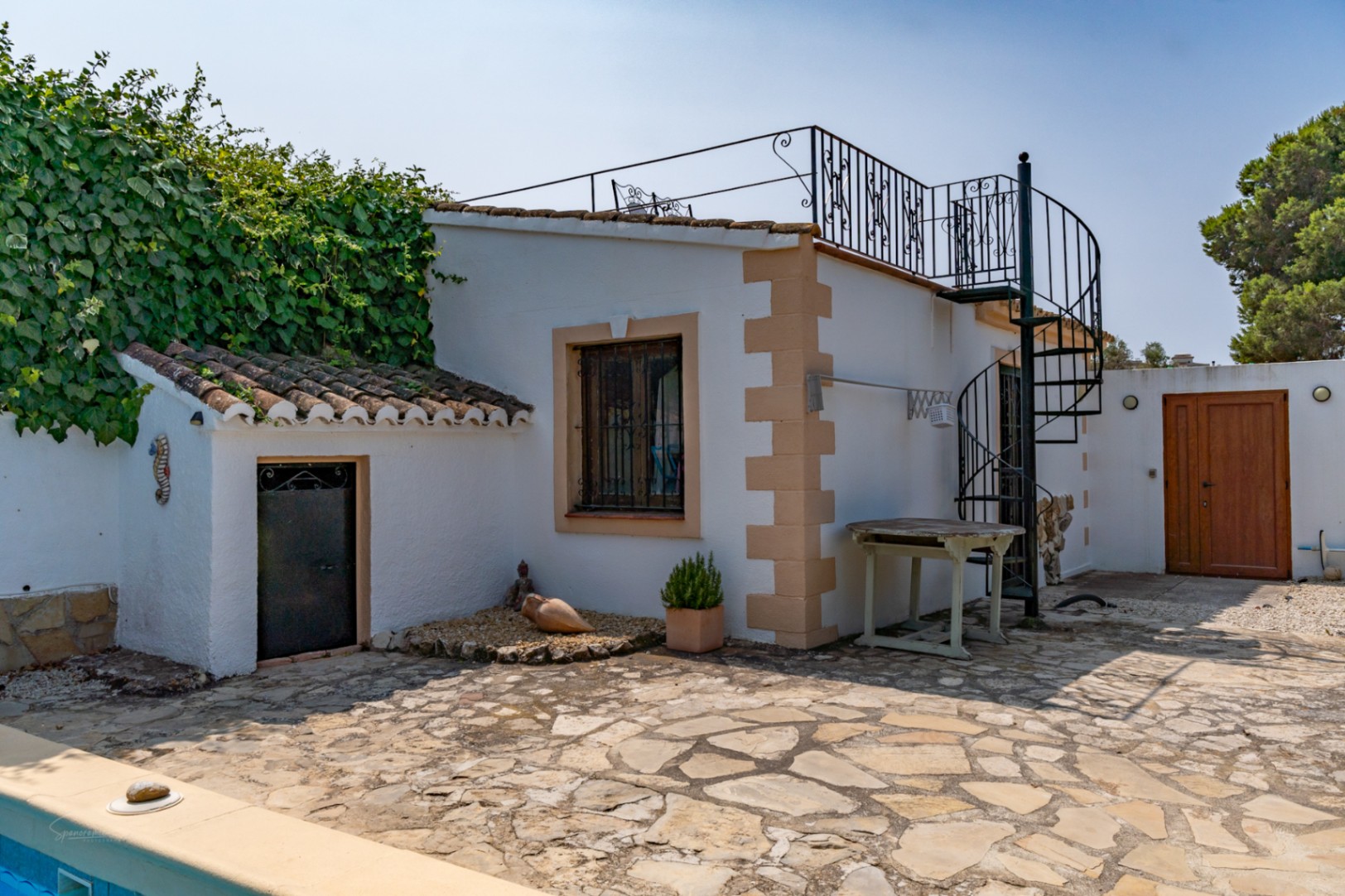 Extérieur d'une maison de campagne avec escalier en colimaçon, annexe avec porte en fer et sol en pierre.