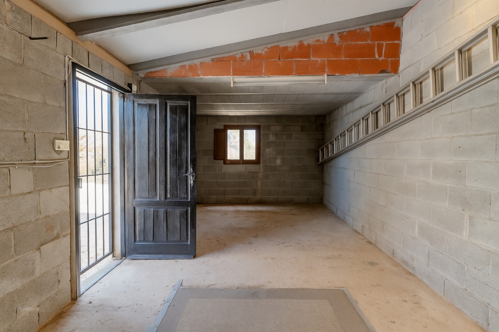 Intérieur de garage avec murs en parpaings, porte en bois ouverte et échelle métallique.