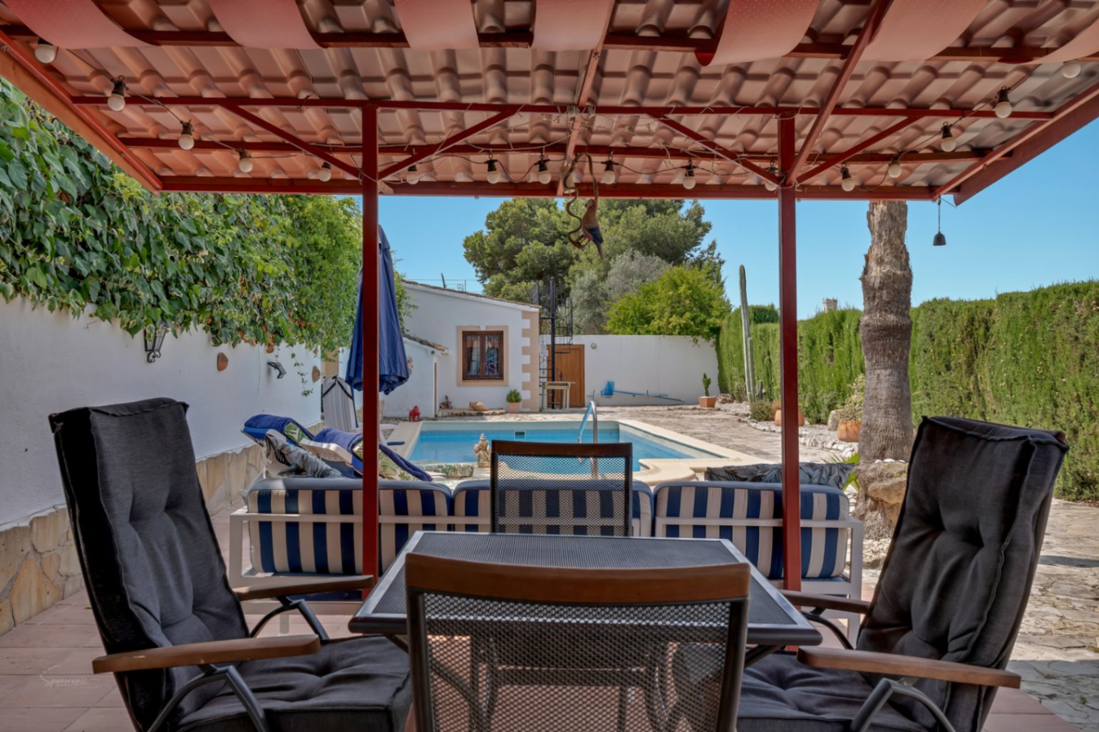 Porche avec vue sur la piscine. Chaises en osier et coussins foncés sous un toit de tuiles rouges.