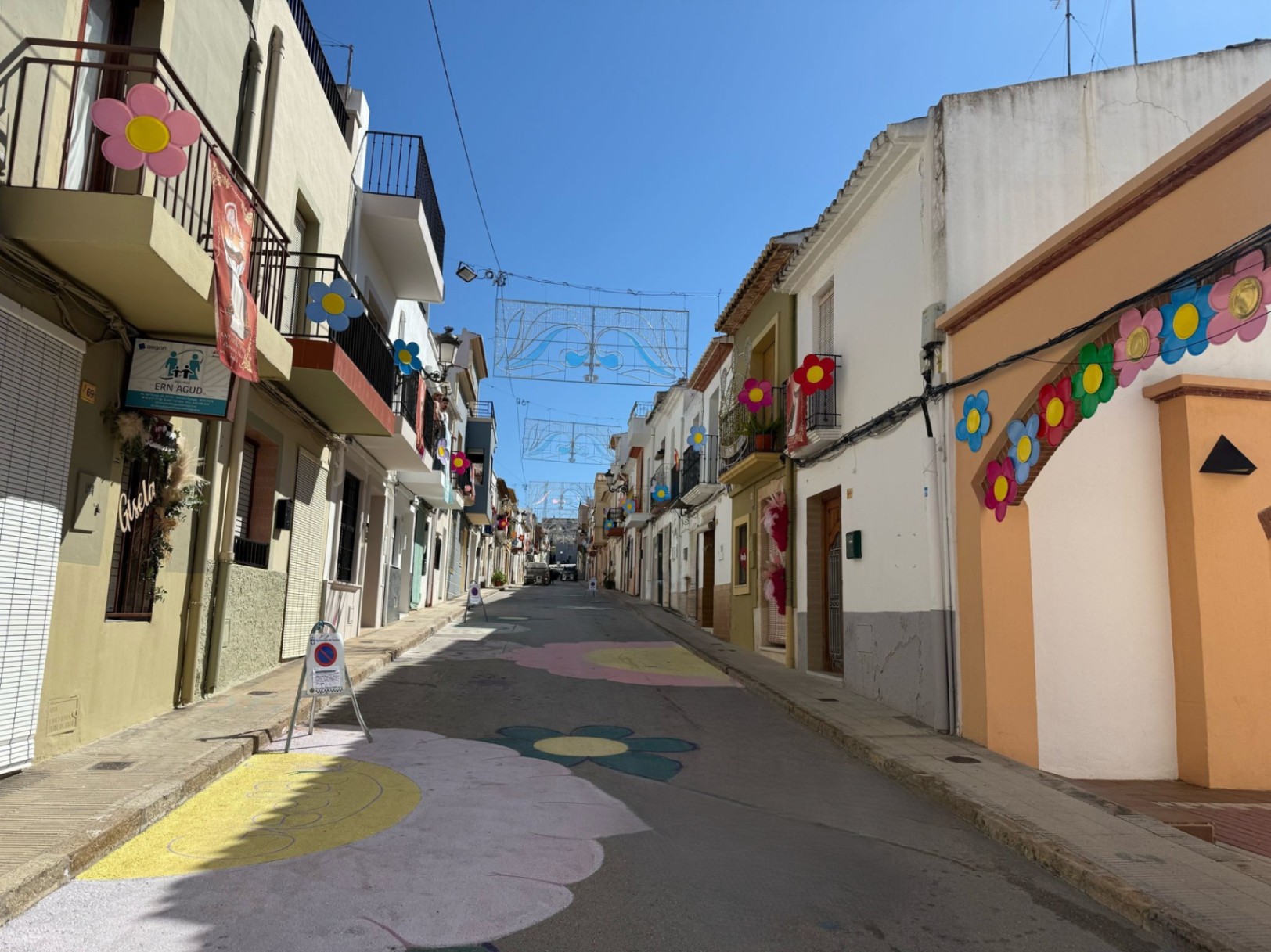 Rue de Teulada décorée de fleurs peintes et d'ornements festifs suspendus.