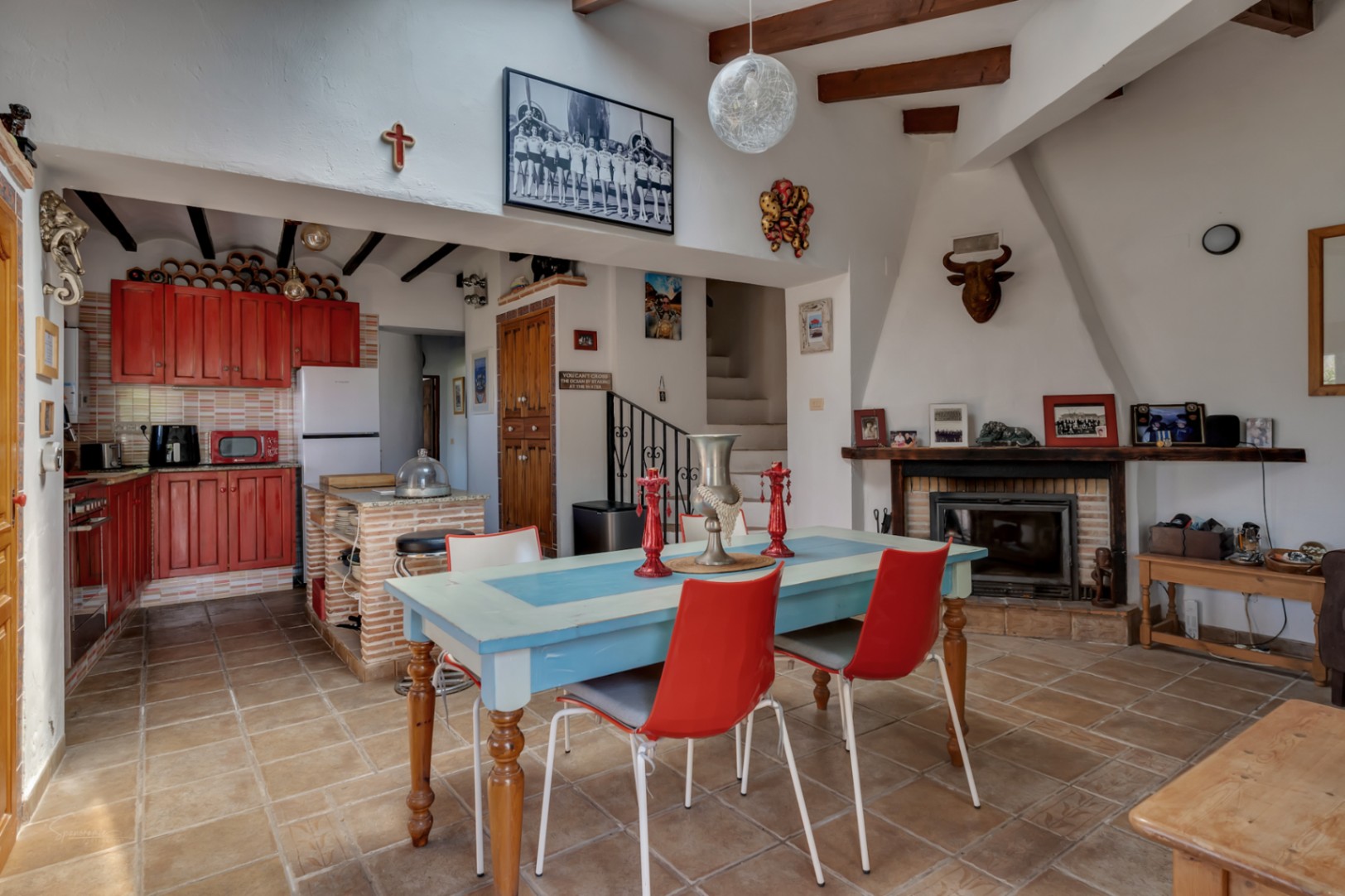 Salle à manger rustique avec table bleue, chaises rouges et cuisine en bois rouge. Cheminée décorée et escalier.